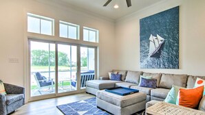 A spacious living room with a large window, a sectional sofa, a coffee table, and a painting of a sailboat.
