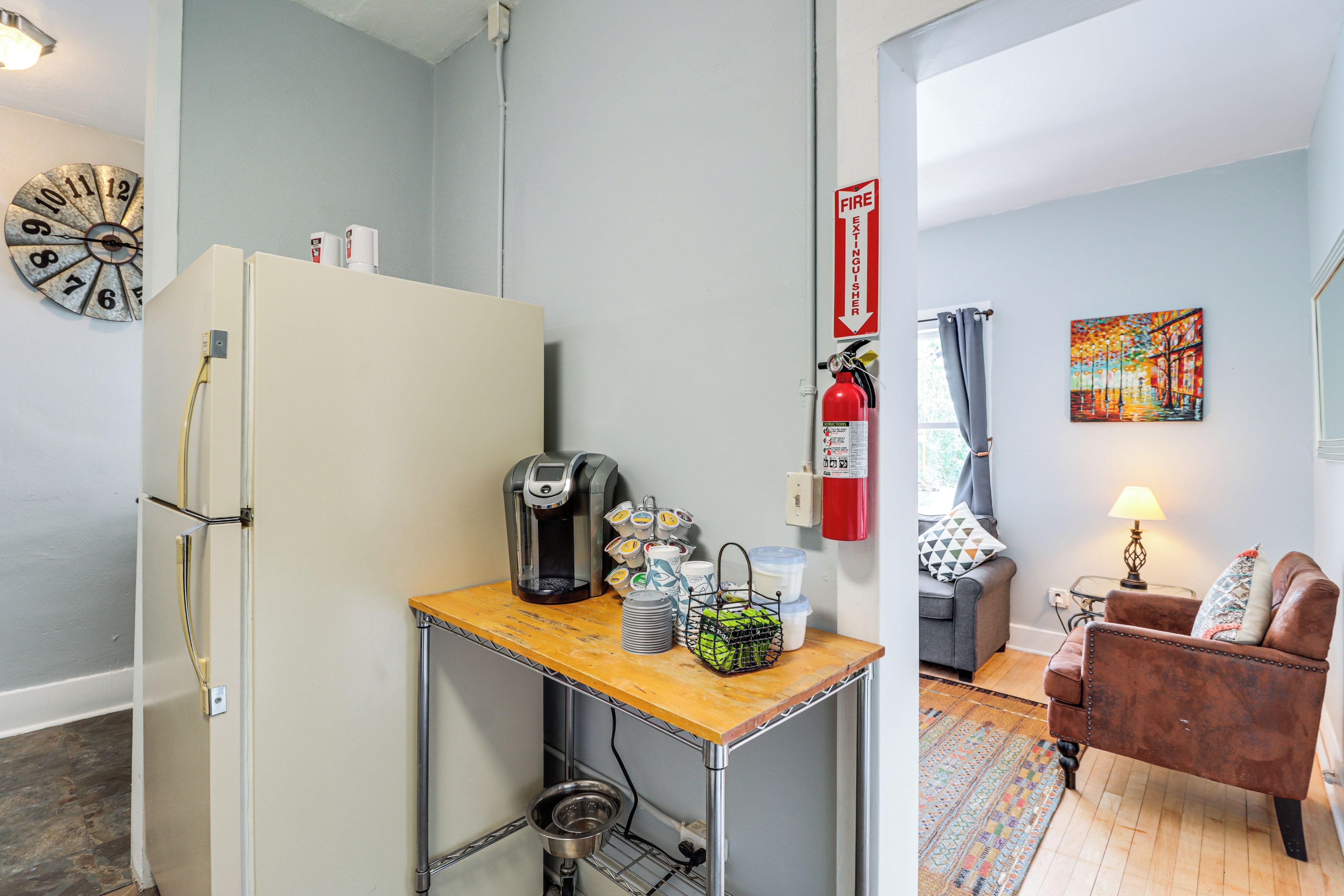 A compact kitchen with a refrigerator, coffee machine, and a small table with a kettle and cups.