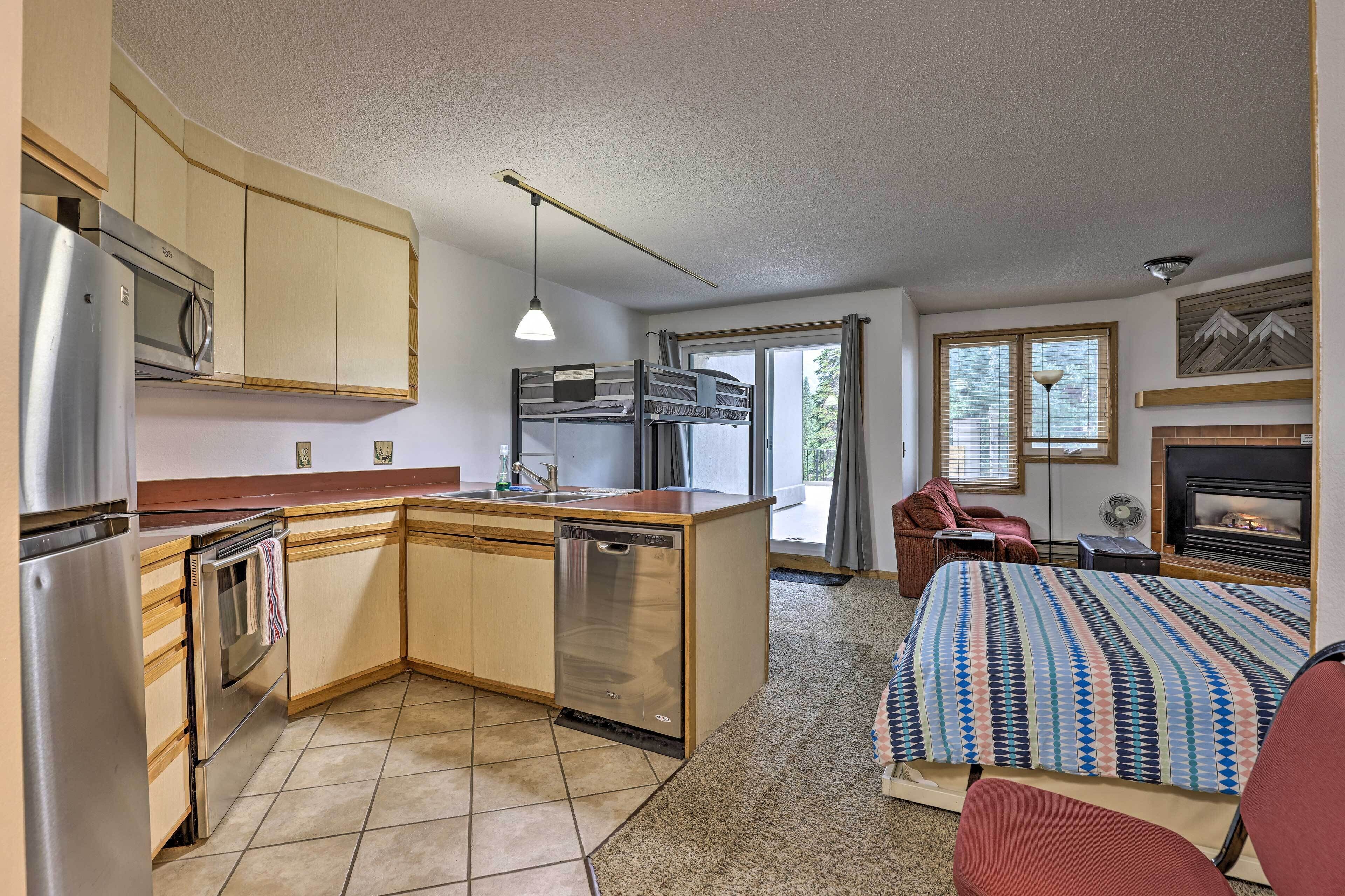 A compact room with a kitchenette, a bed with a patterned blanket, a fireplace, and a window with curtains.