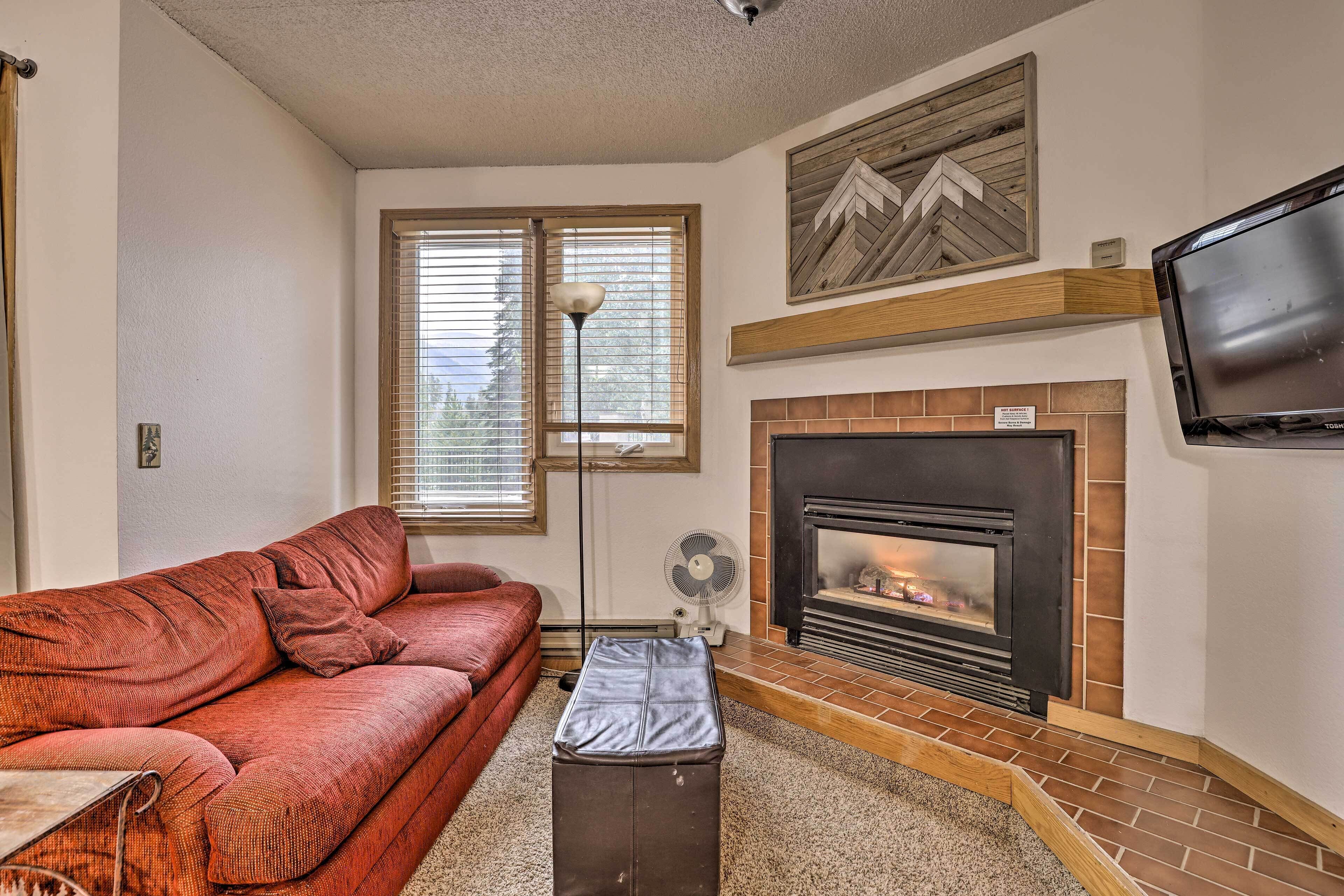 A living room with a red sofa, a fireplace, a TV mounted on the wall, and a window with blinds.