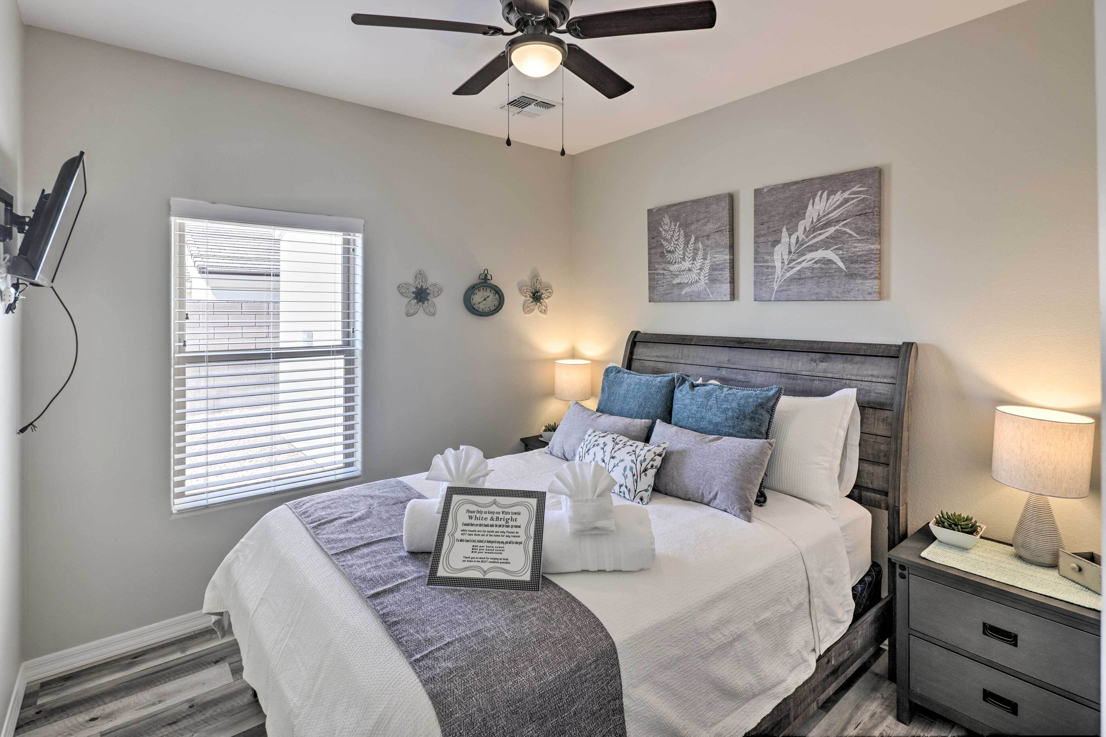 A bedroom with a bed, a ceiling fan, a window with blinds, a bedside table with a lamp, and wall art.