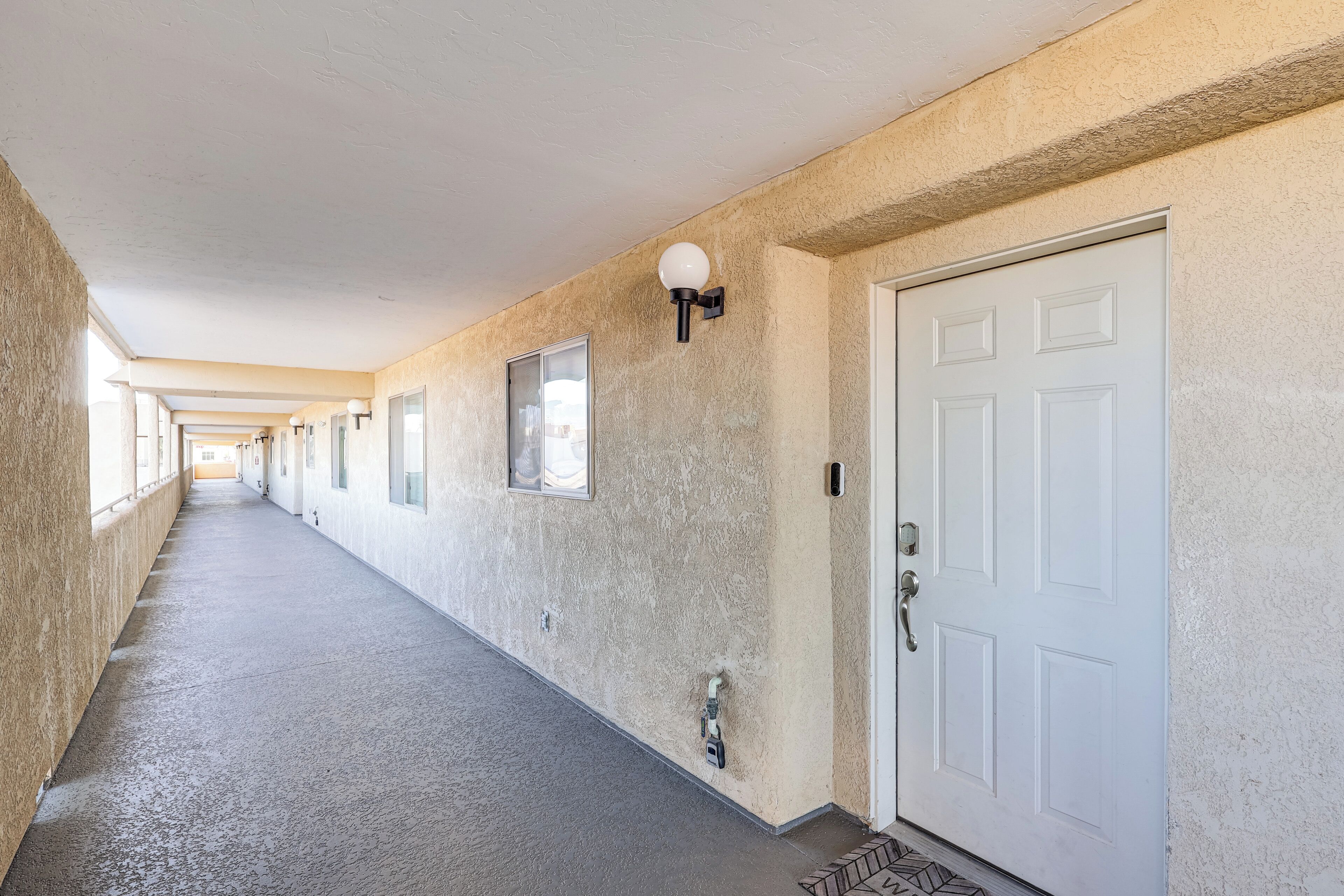 A long, narrow hallway with a white door on the right, a window on the left, and a wall-mounted light fixture.