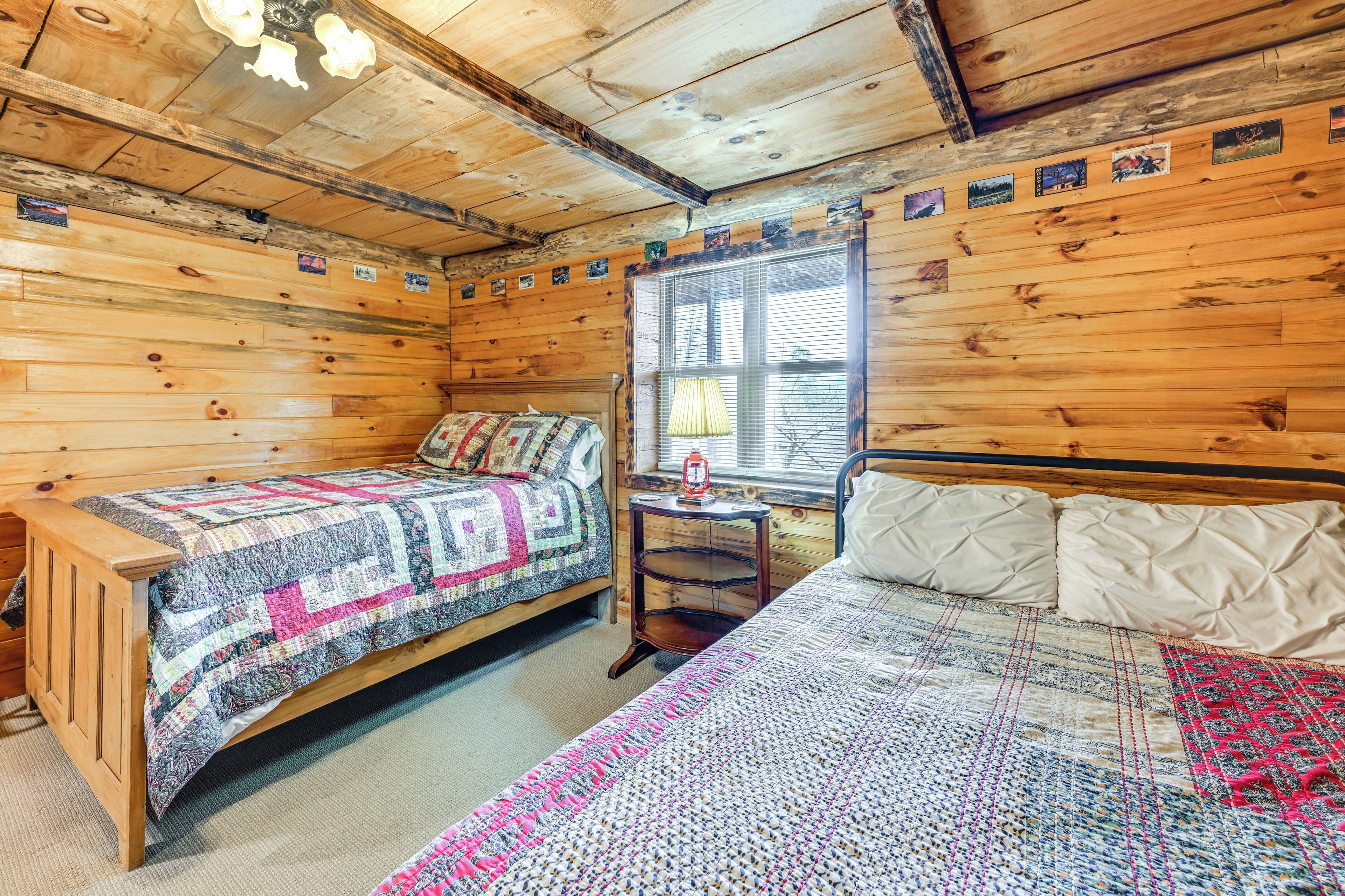 A wooden cabin room with two beds, a window with blinds, and a small table with a lamp.