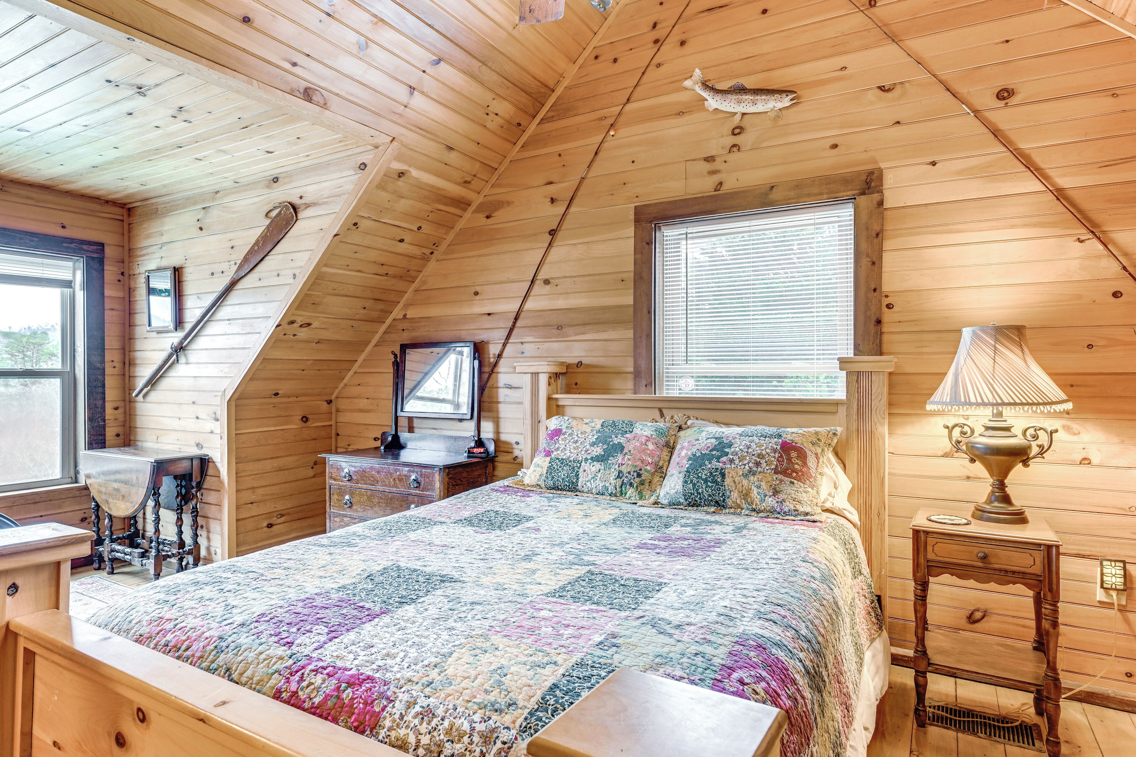 Une chambre confortable avec un plafond en bois, un lit recouvert d’une courtepointe aux couleurs vives, une table de chevet avec une lampe et une fenêtre équipée de stores.