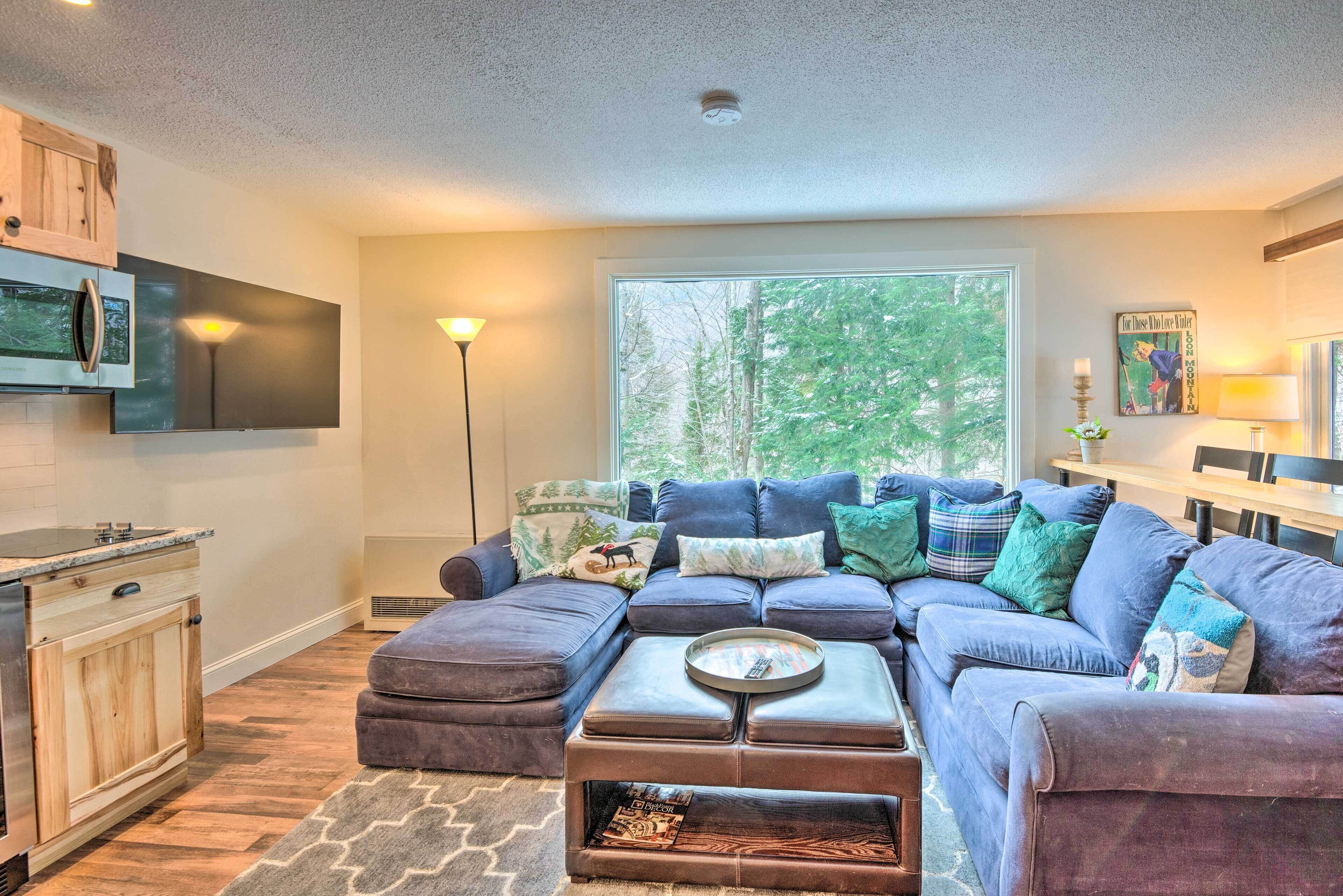 A modern living room with a sectional sofa, a flat-screen TV mounted above the fireplace, and a large window offering a view of trees.