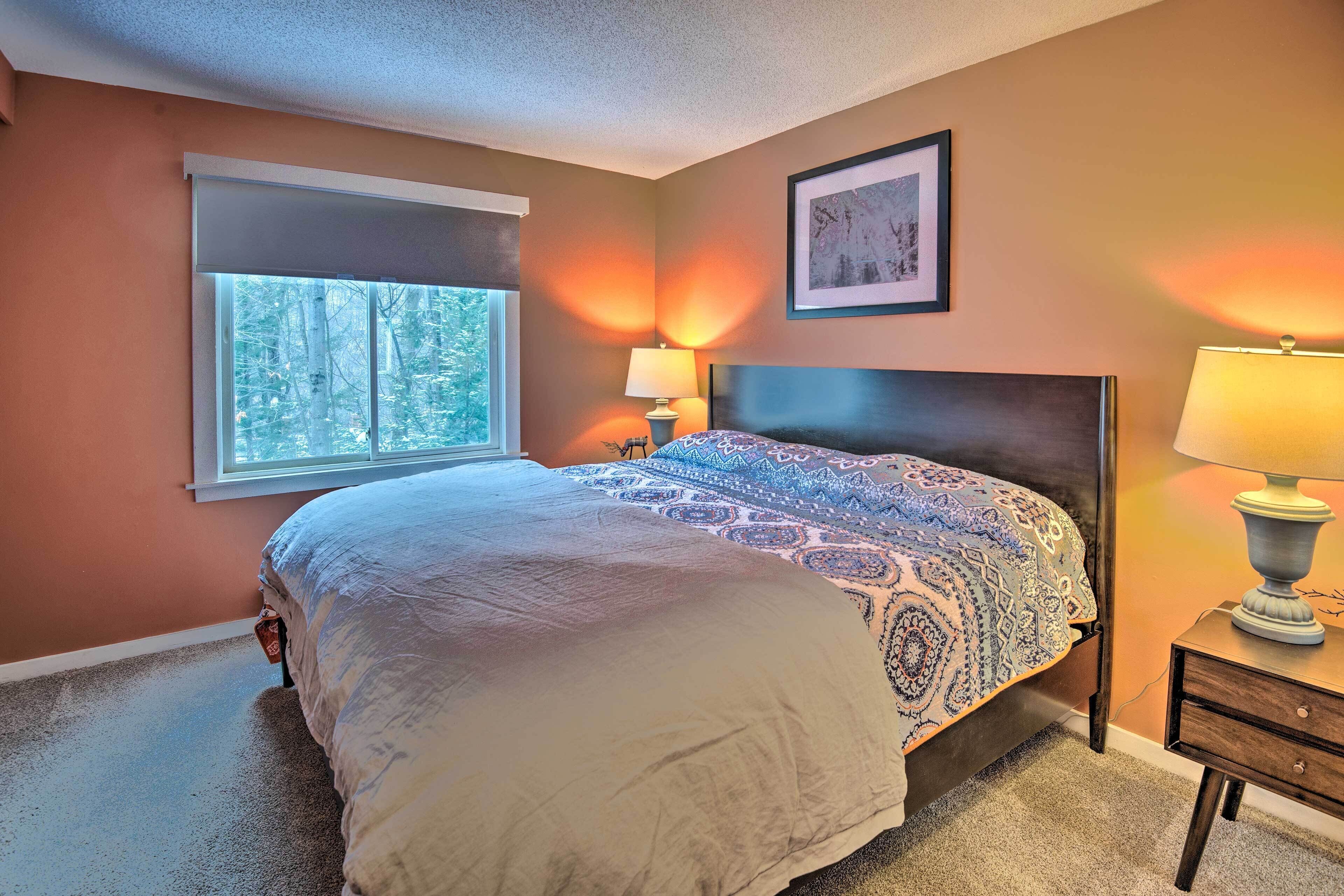 A bedroom with a large bed, two bedside lamps, a window with a view of trees, and a framed picture on the wall.