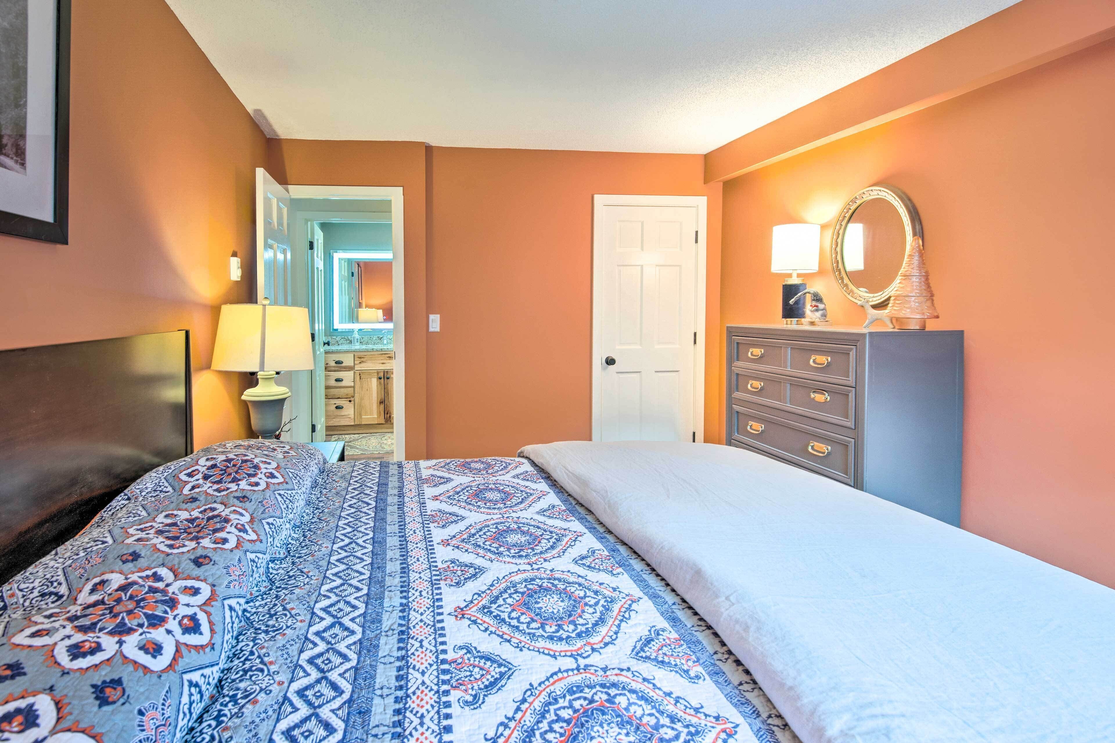 Une chambre à coucher avec une couvre-lit à motifs, une commode avec des tiroirs, un miroir et une porte donnant sur une autre pièce.