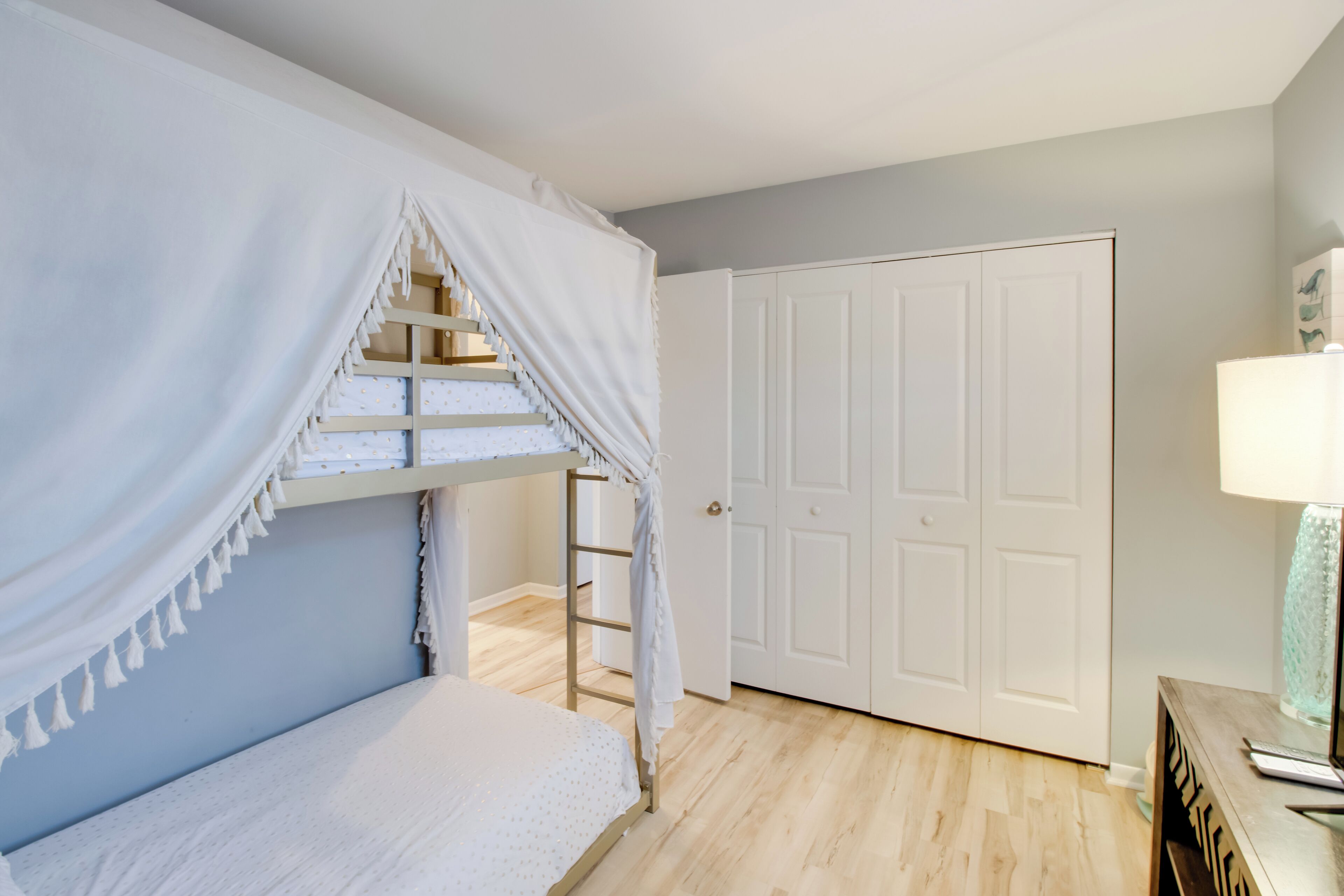 A bedroom with a bunk bed, a ladder, a white door, and a bedside table with a lamp.