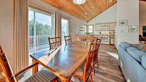Ruime eetruimte met een houten tafel en stoelen, een keuken op de achtergrond, en een balkon met uitzicht op bomen.