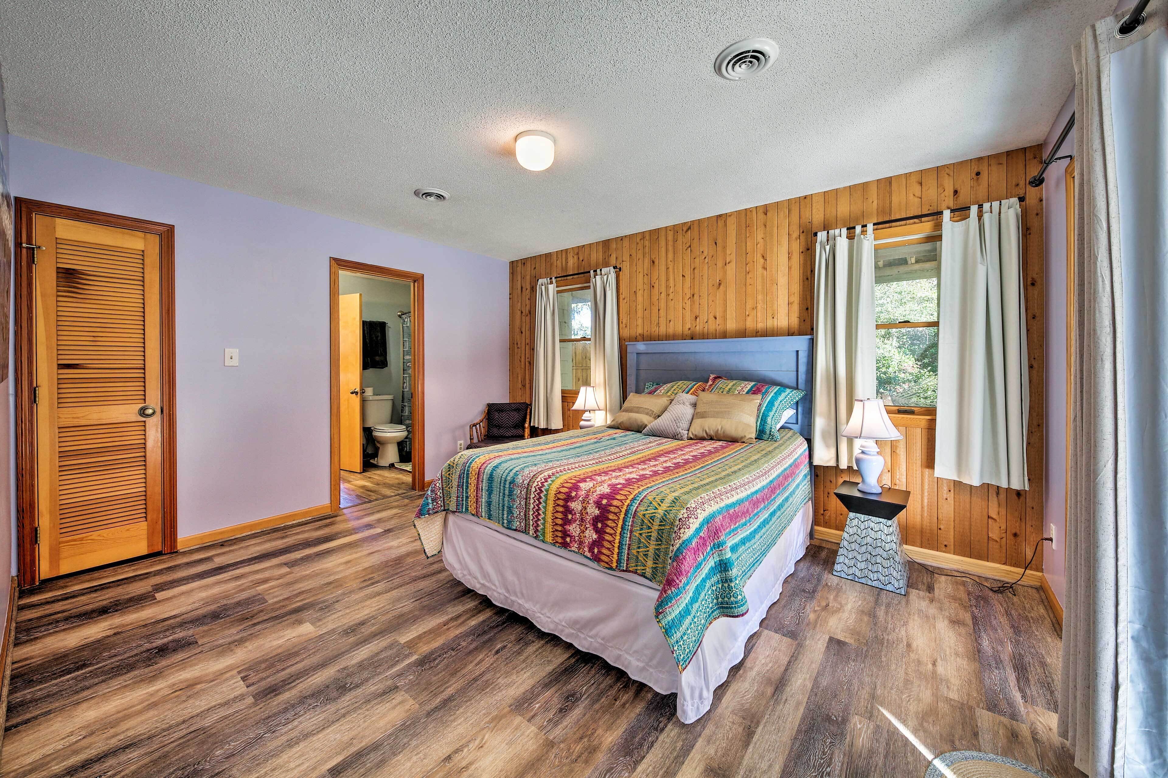 A bedroom with a wooden headboard, a large bed with a colorful blanket, a nightstand with a lamp, and a window with curtains.