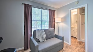 Una habitación con un sofá gris, una lámpara, una ventana con cortinas y una puerta que da a un baño.