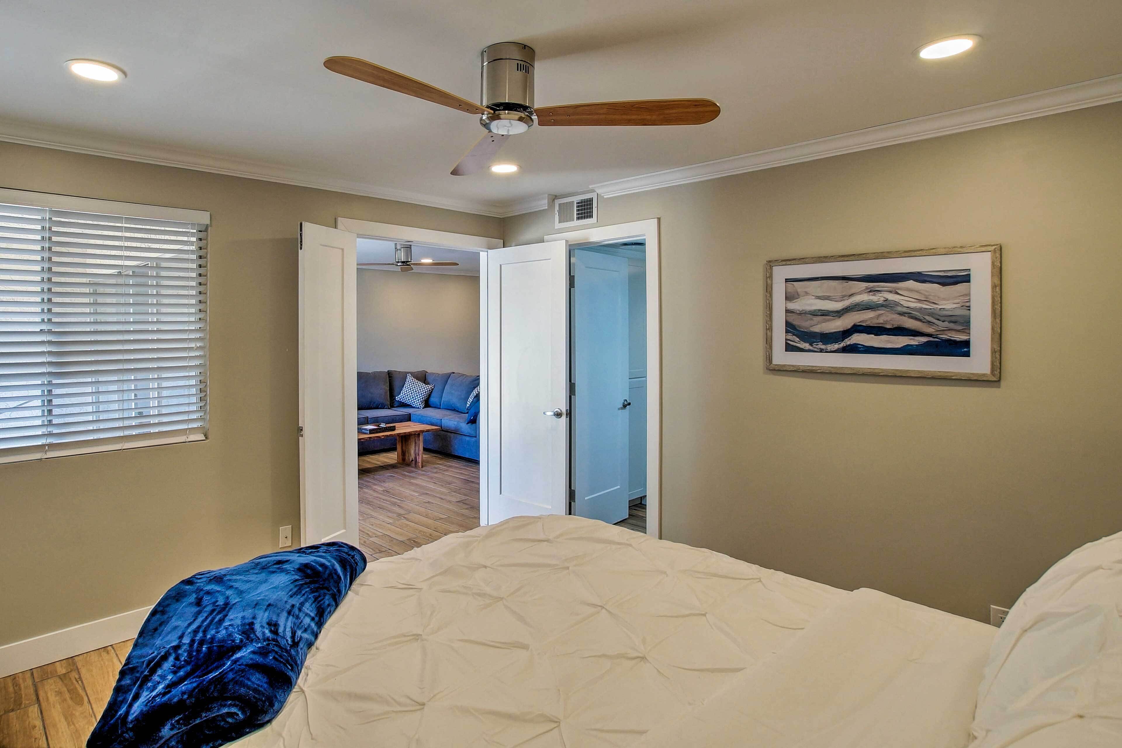 A bedroom with a bed, a ceiling fan, a painting, and a door leading to another room.