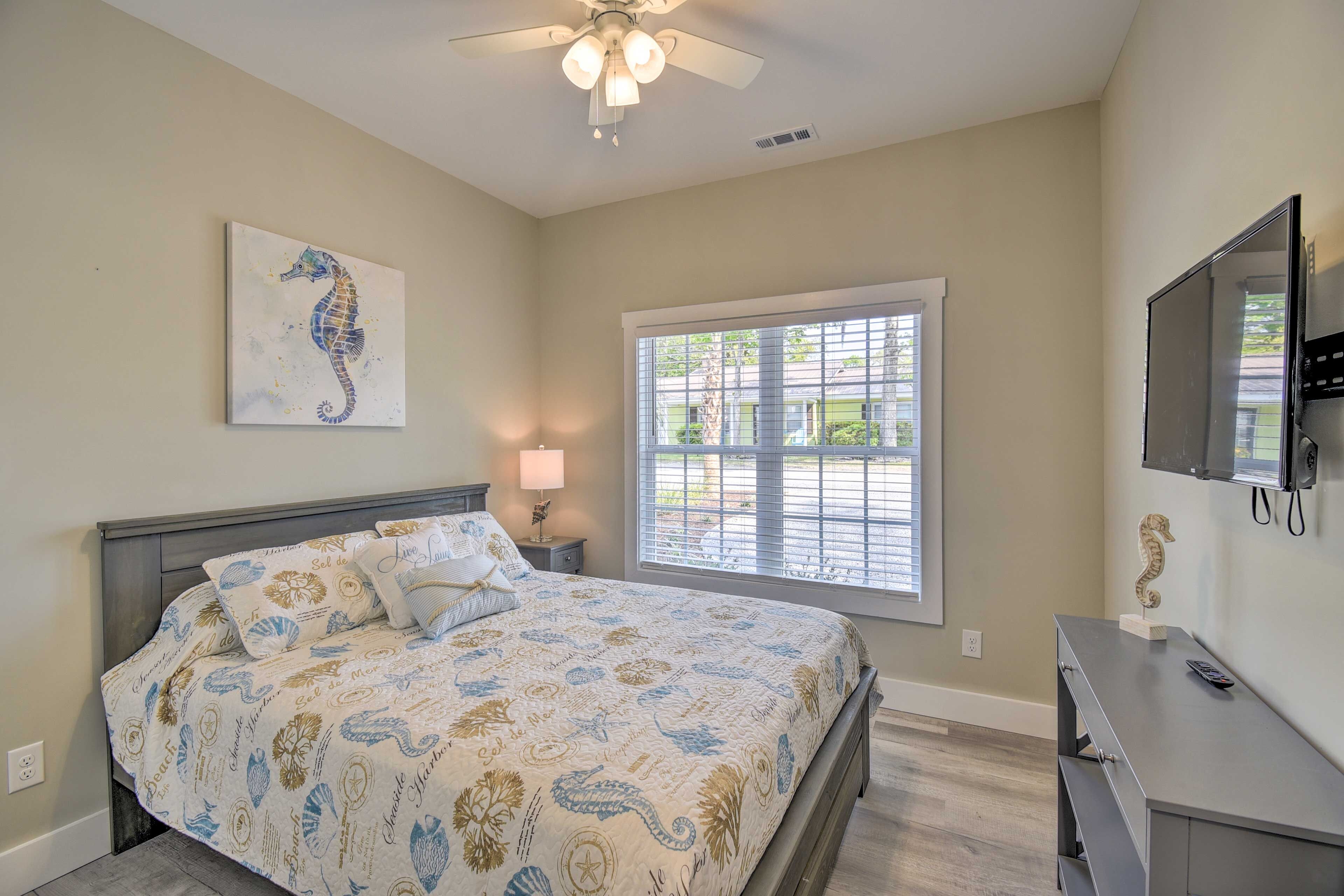 A bedroom with a bed, a ceiling fan, a window with blinds, a TV mounted on the wall, and a small table with a lamp.