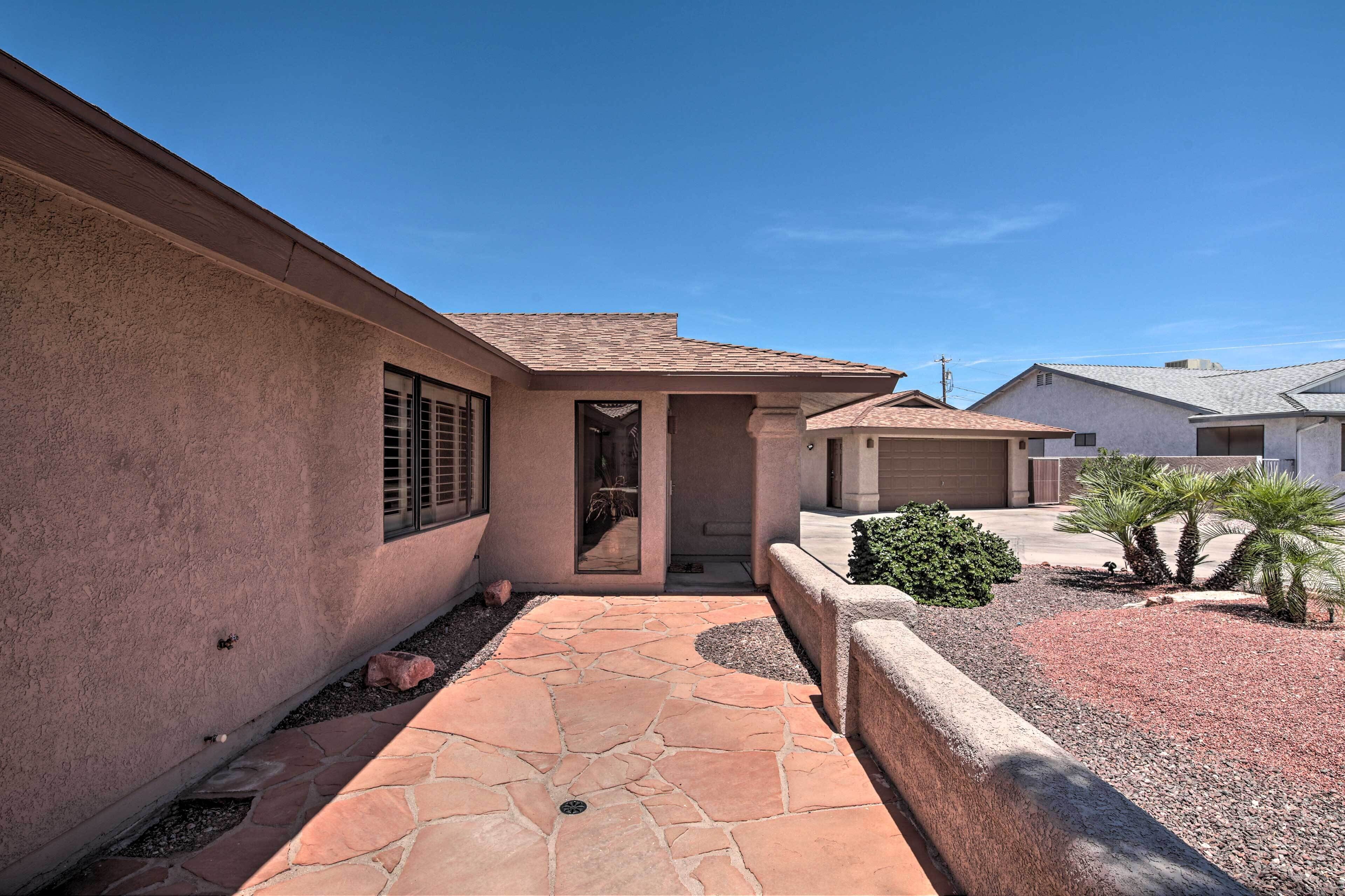 A residential property with a stone pathway leading to a door, a small garden with shrubs and a palm tree, and a neighboring house with a garage.