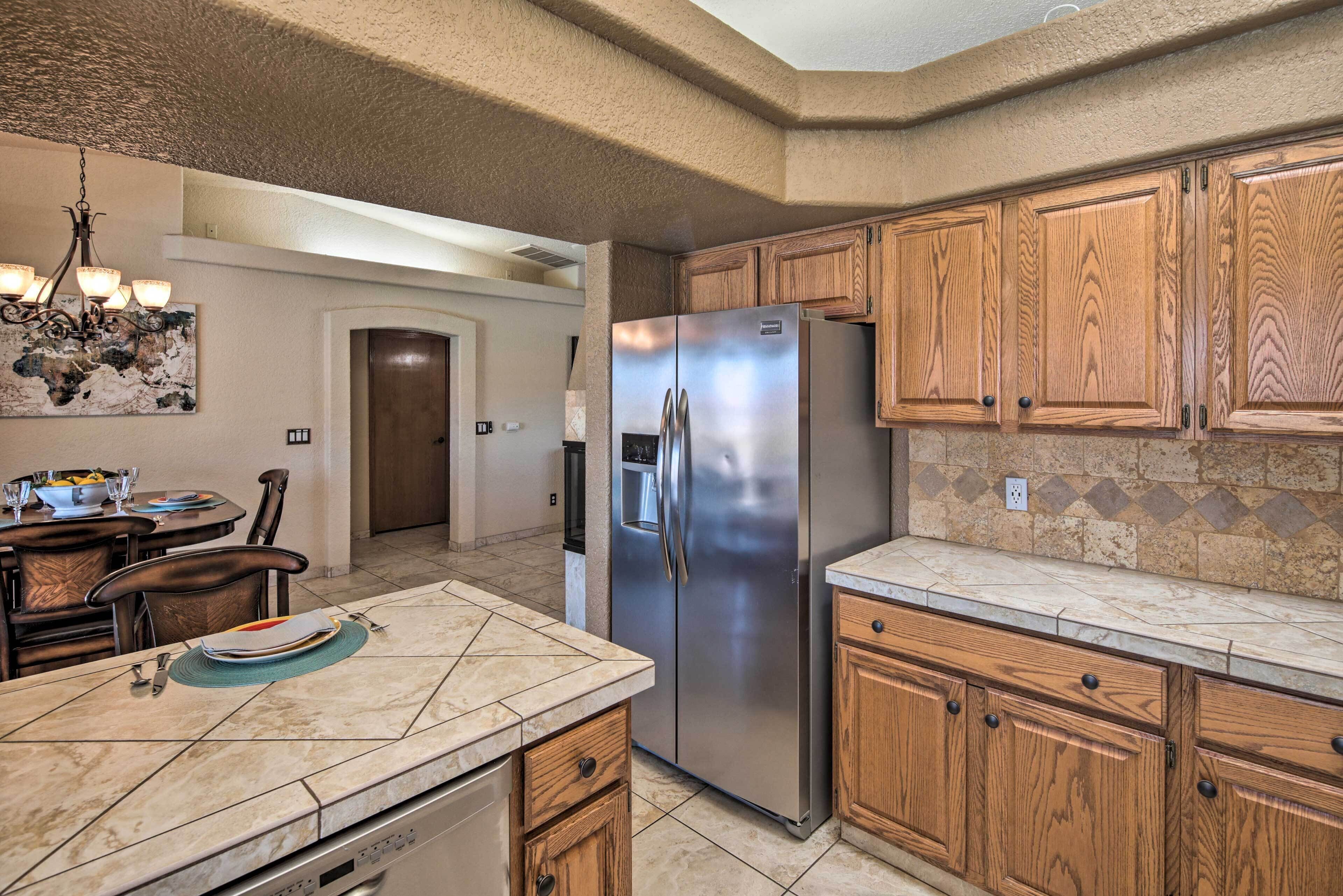 A kitchen with a stainless steel refrigerator, wooden cabinets, a marble countertop, and a dining area with chairs and a table.