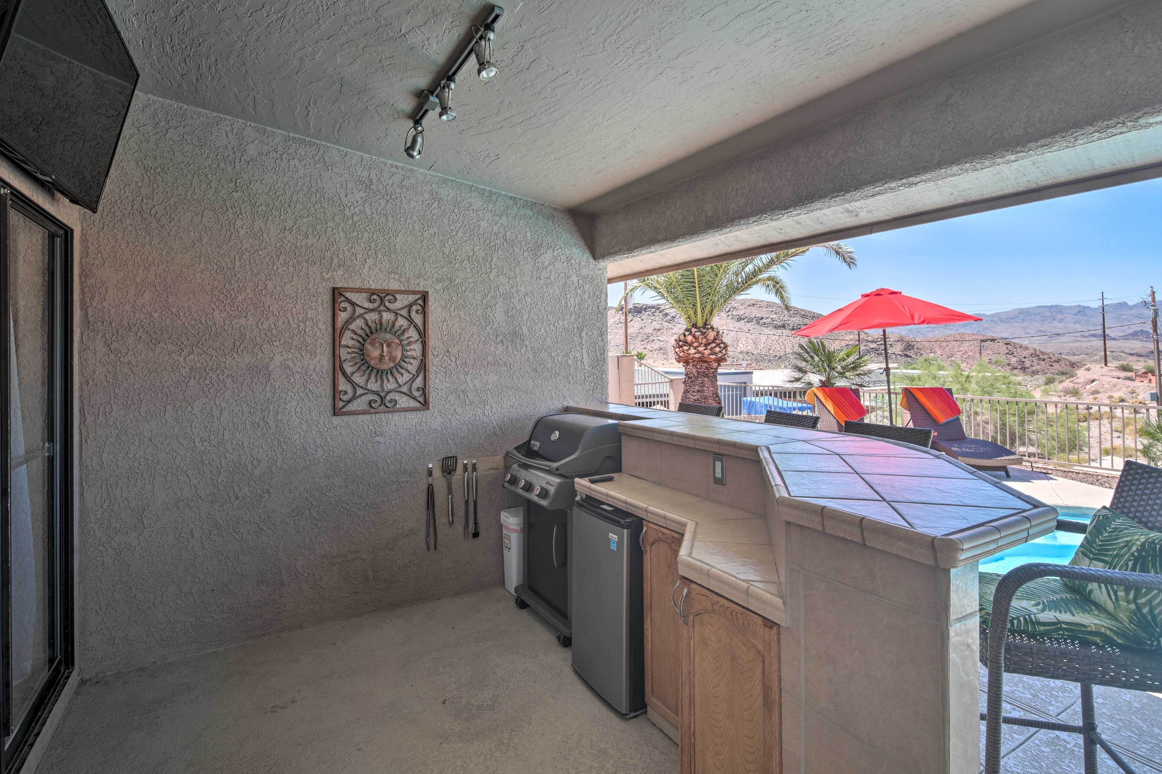 An outdoor kitchen with a grill, refrigerator, and bar area, overlooking a pool and desert landscape.