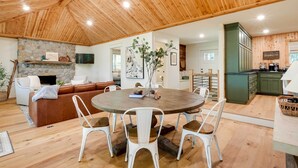A cozy living room with a stone fireplace, a round dining table with white chairs, a brown leather sofa, and a kitchen area with green cabinets.