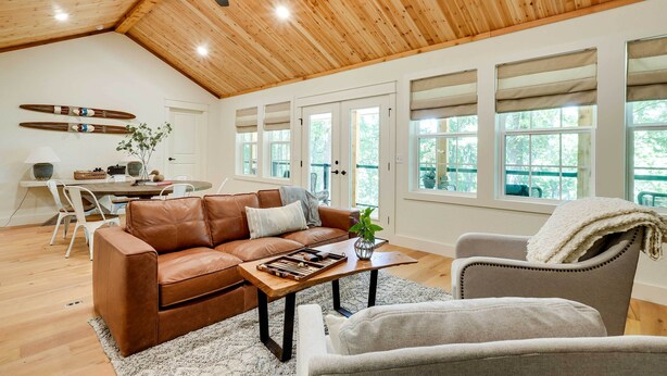 A spacious living room with a high wooden ceiling, a brown leather sofa, a grey armchair, and a dining area with a white table and chairs.