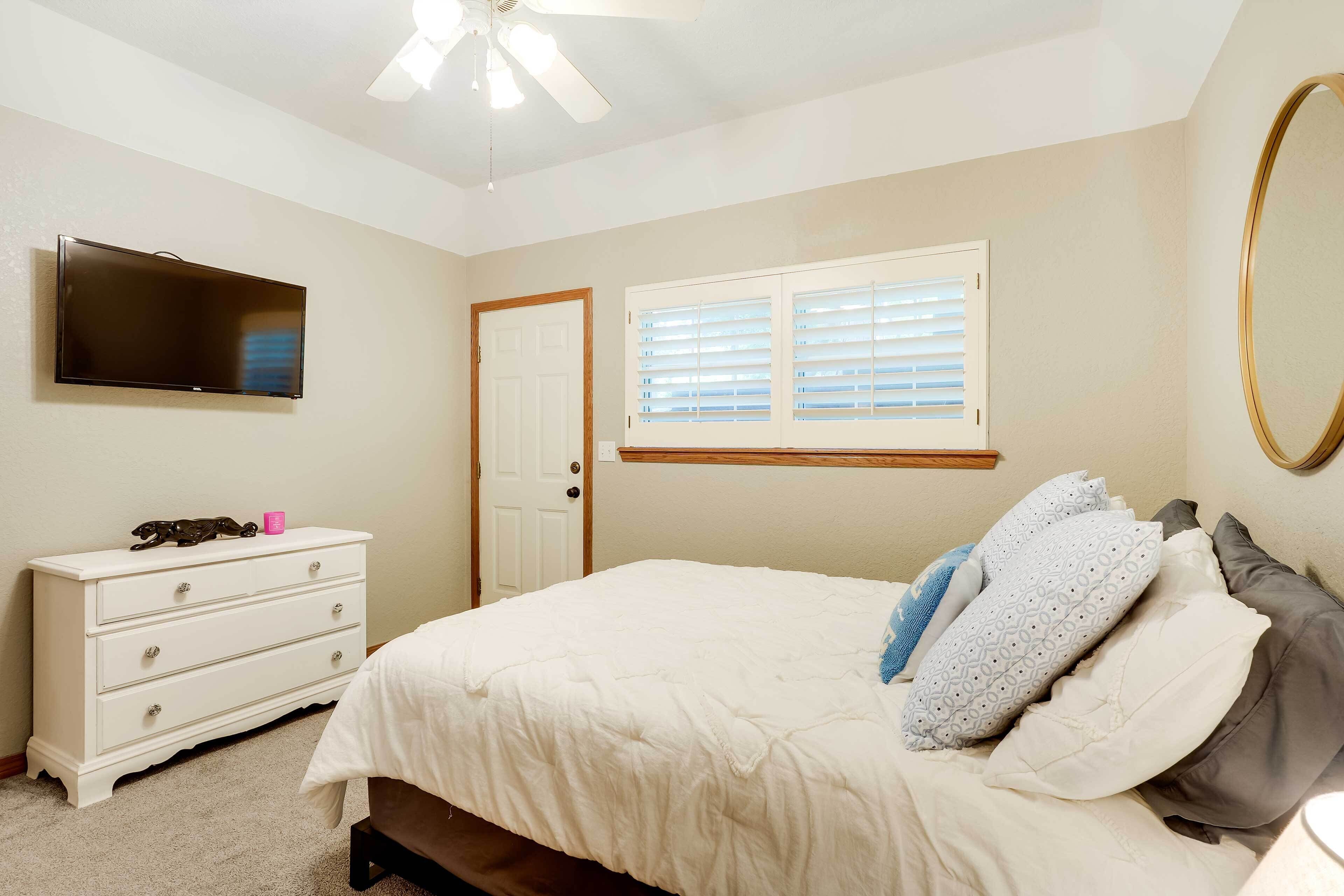 A bedroom with a bed, a dresser, a television, a window with blinds, and a ceiling fan.