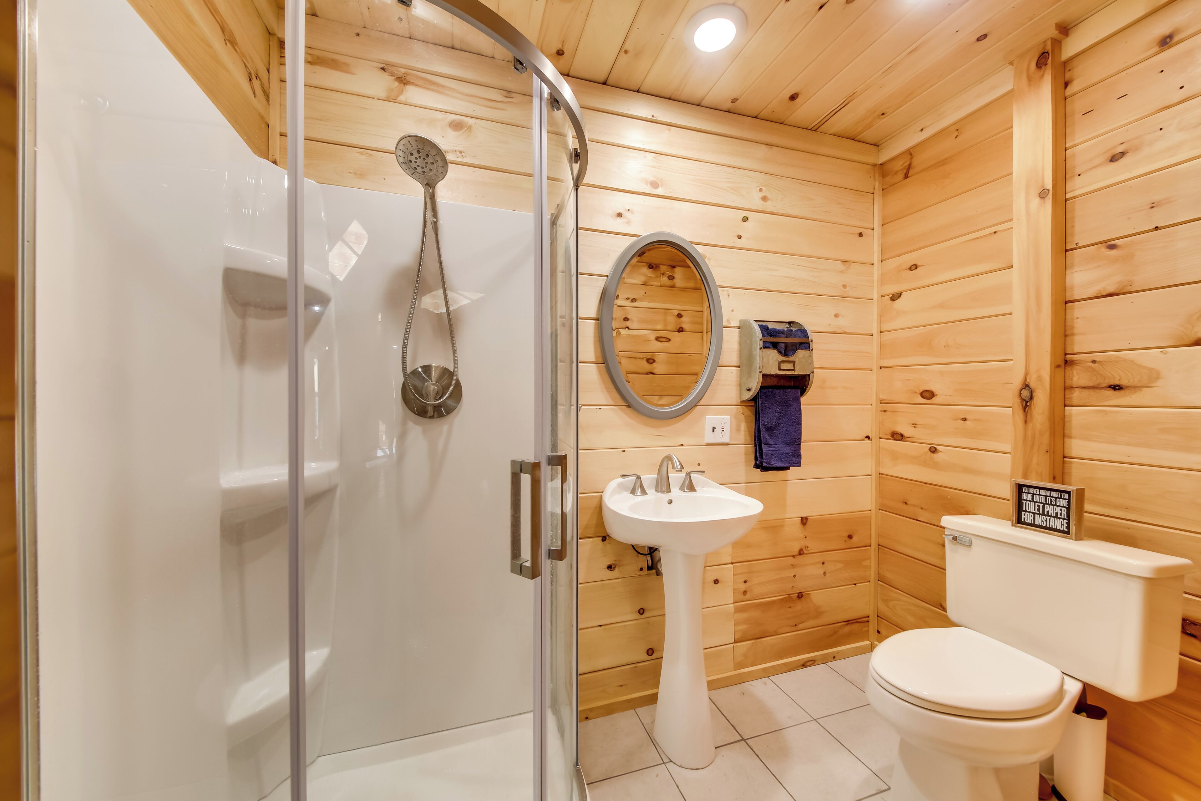 Baño de cabaña de madera con mampara de vidrio, lavabo pedestal, inodoro y pared de madera.