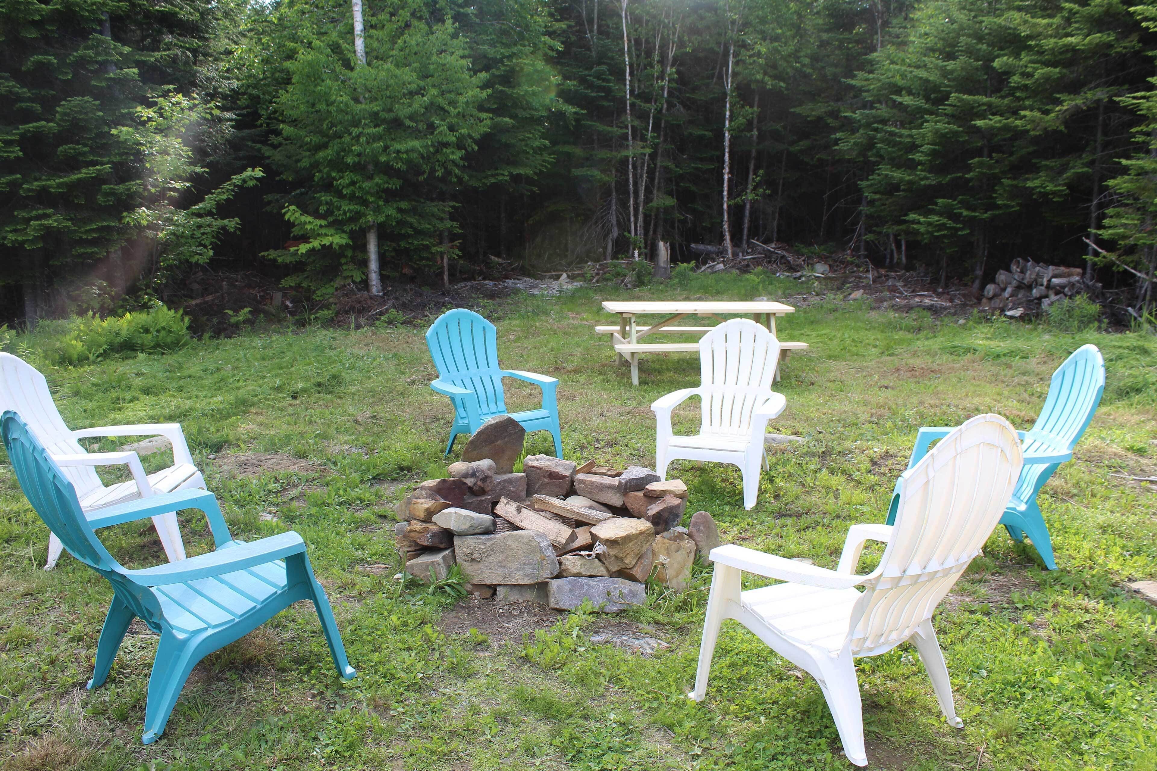 A group of Adirondack chairs arranged around a wooden fire pit in a forested area.