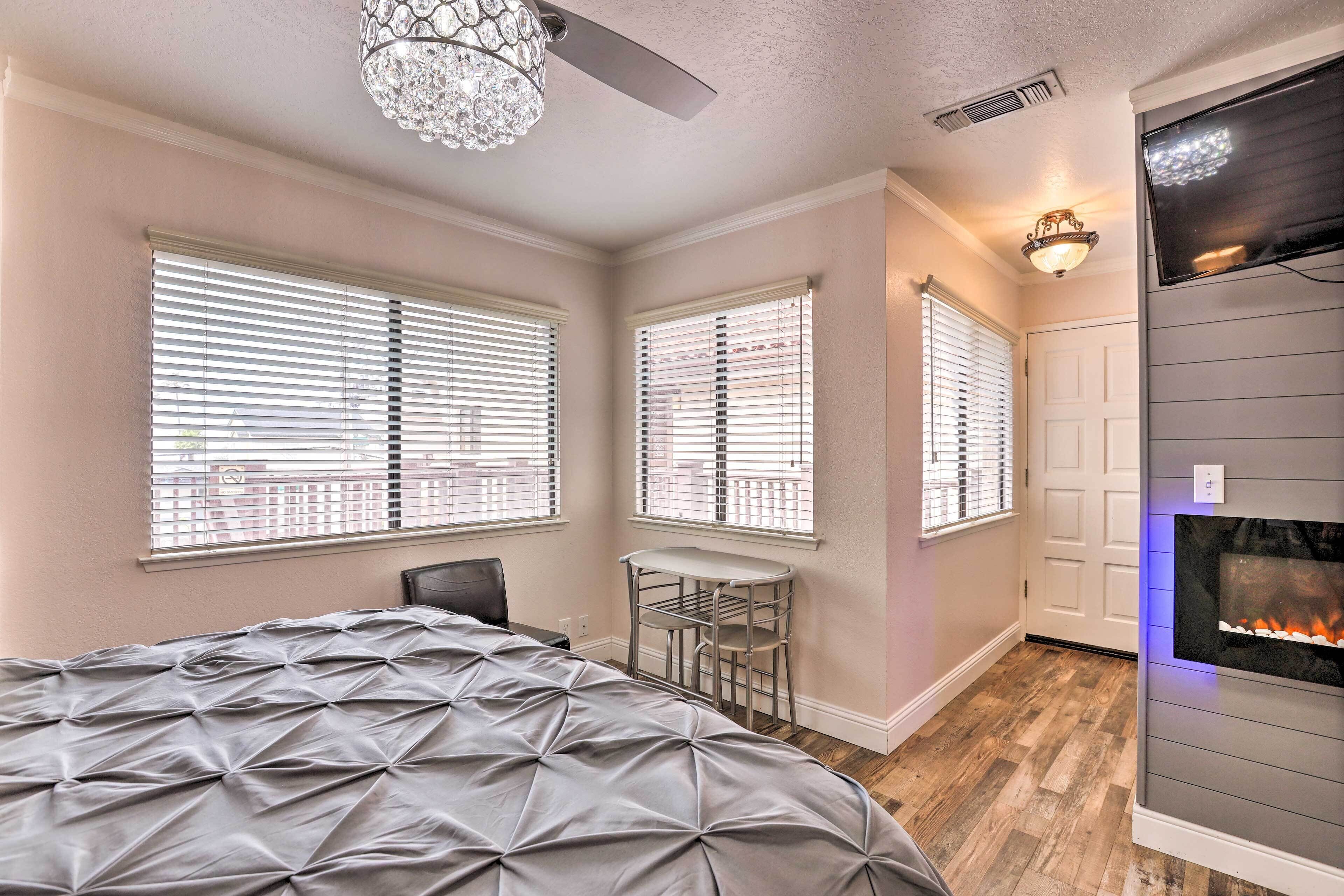 A bedroom with a bed, a chair, a small table, a fireplace, and large windows with blinds.
