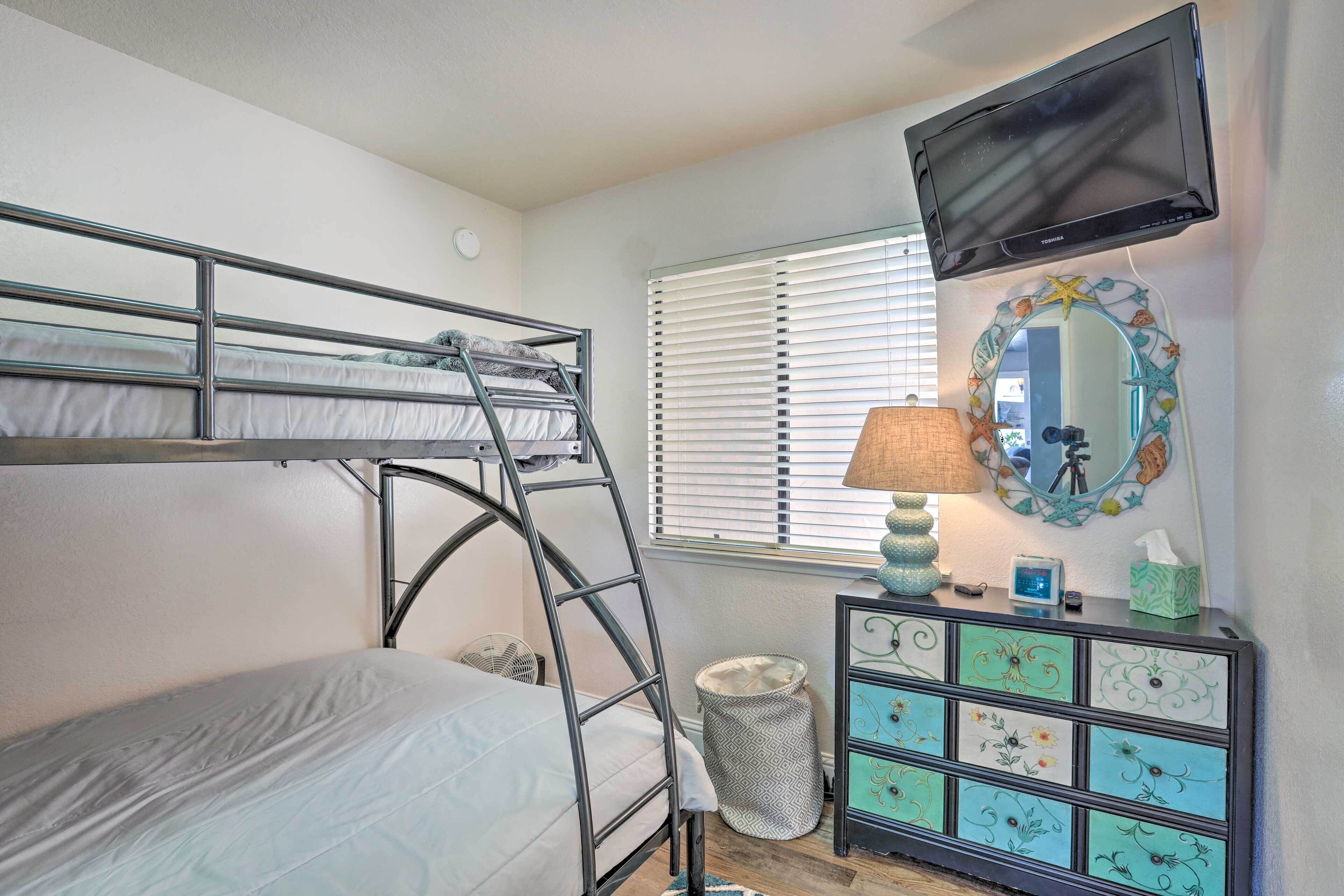 A bunk bed with a ladder, a television mounted on the wall, a dresser with decorative panels, and a window with blinds.