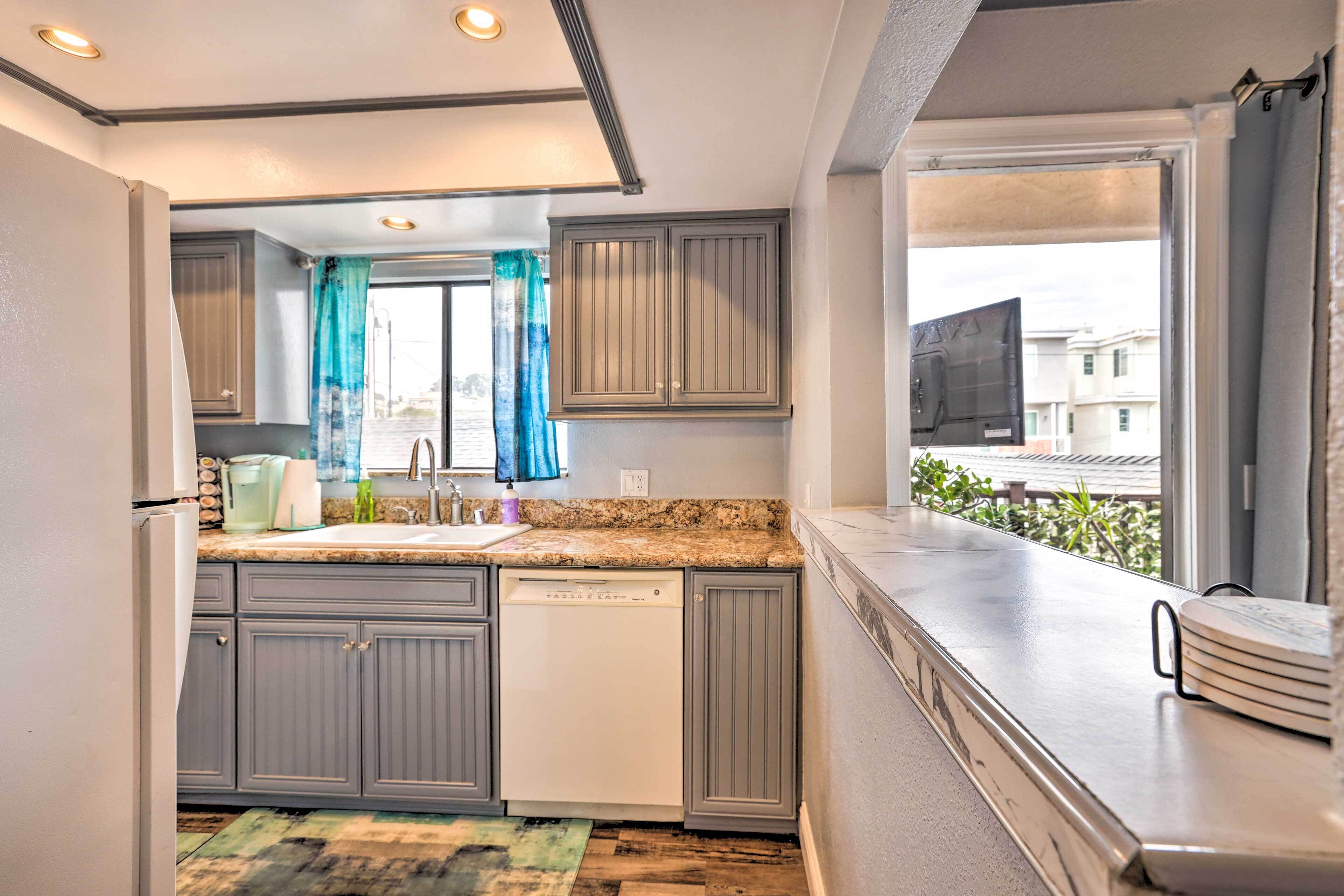 A modern kitchen with grey cabinets, a dishwasher, and a window with a view of buildings and greenery.