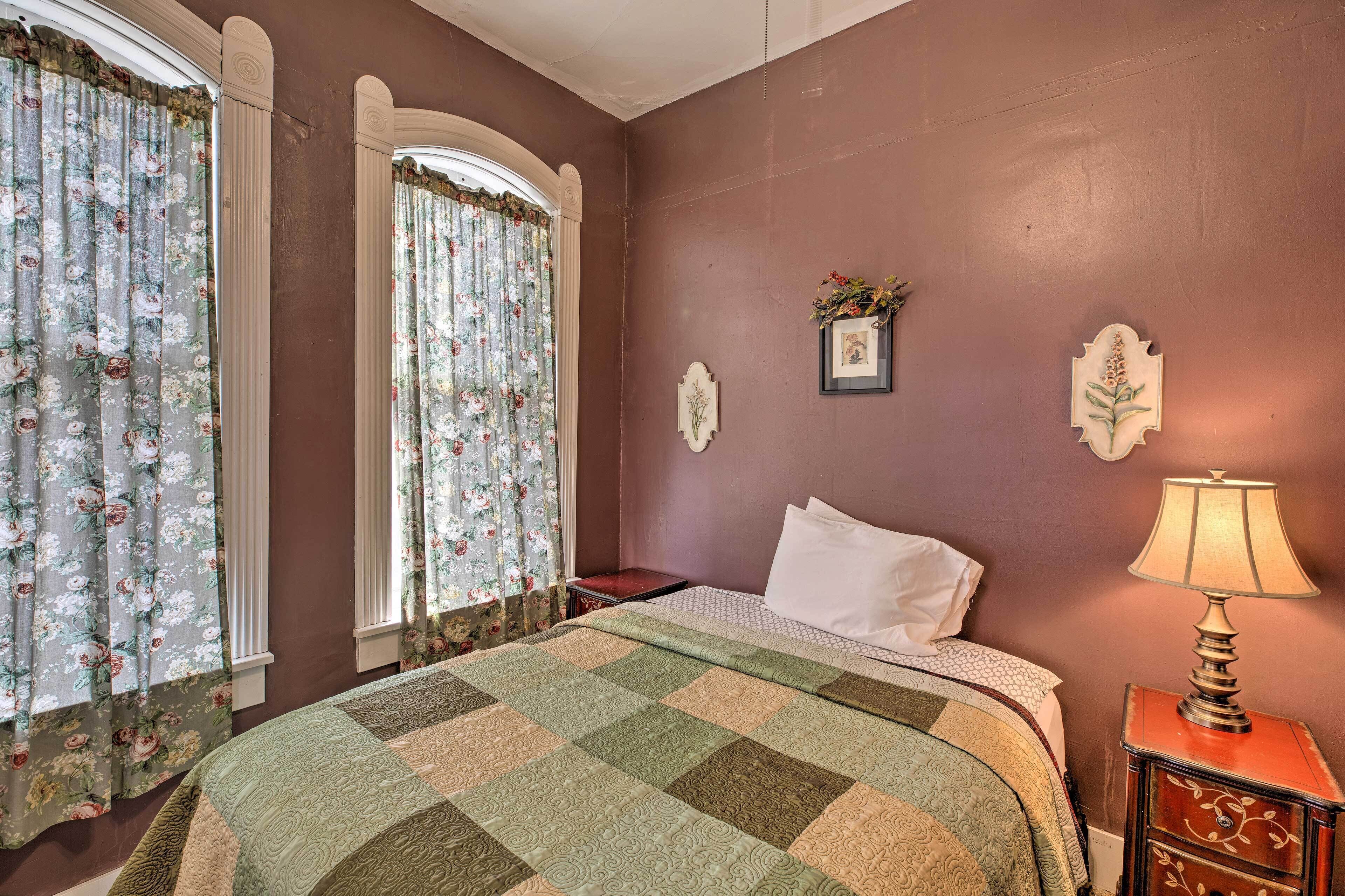 Une chambre à coucher avec une couvre-lit à carreaux, des rideaux à motifs floraux, une lampe de chevet et un tableau encadré accroché au mur.