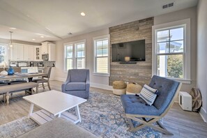A spacious living room with a fireplace, large windows, and a kitchen in the background. - Kure Beach Townhome: Short Walk to the Beach! (Kure Beach)