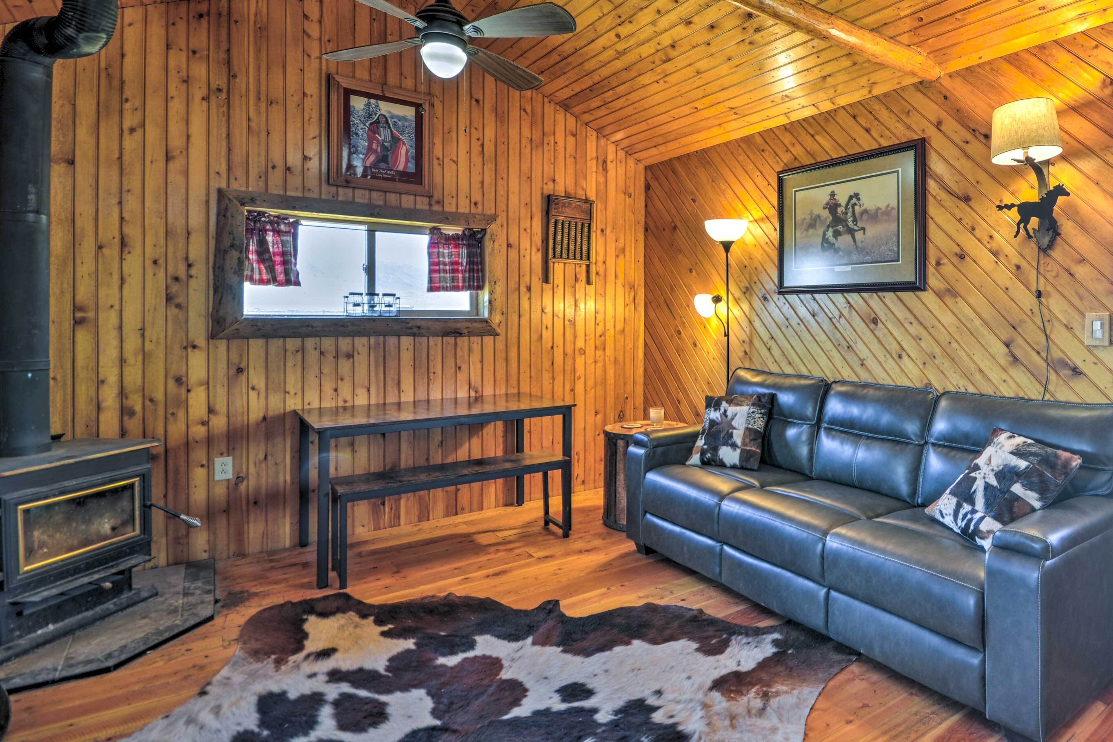 Interior acogedor de una cabaña con sofá de cuero, paredes de madera, chimenea y un ventanal con cortinas.