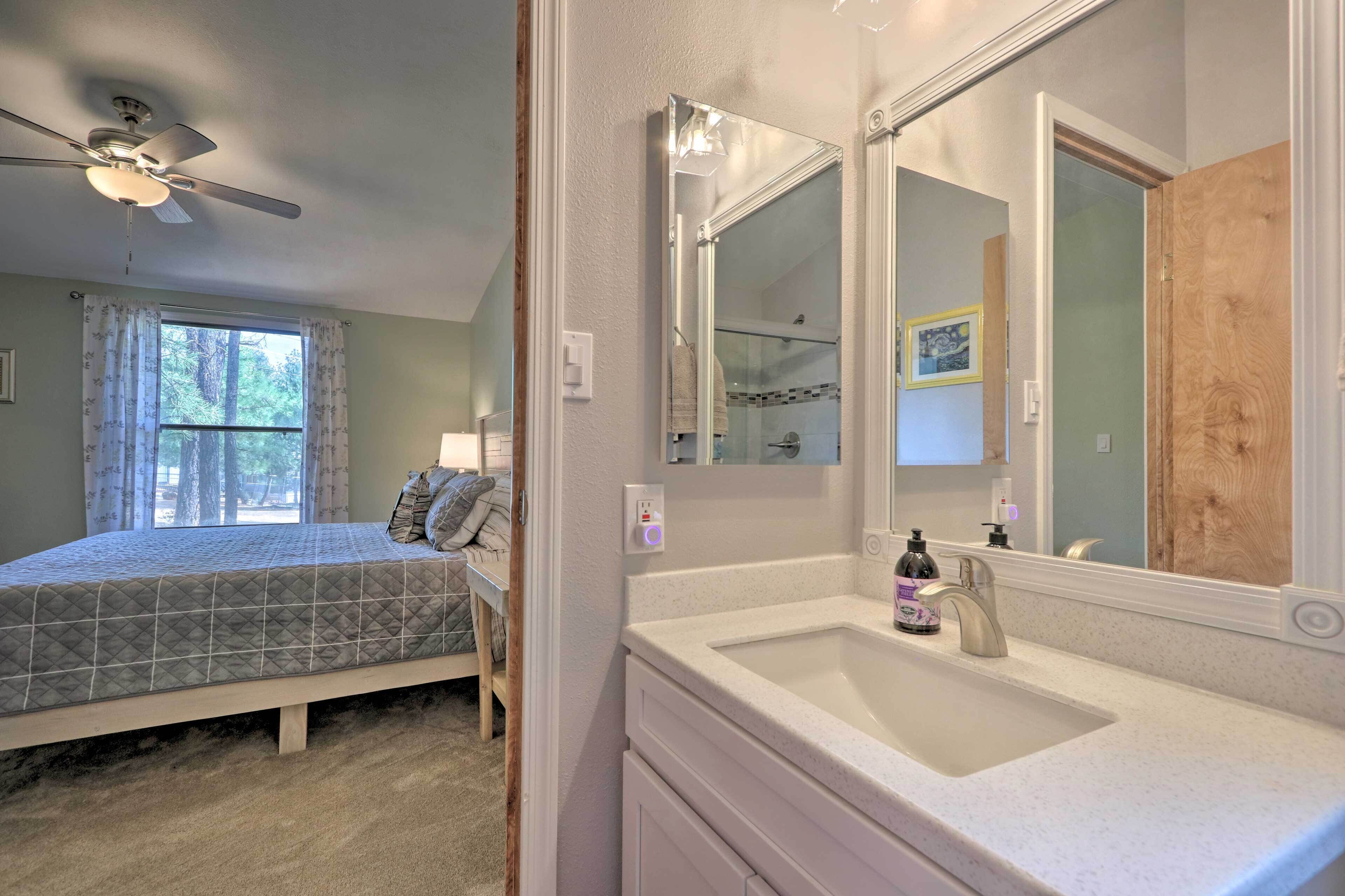 A bedroom with a bed, a ceiling fan, a window with curtains, and a bathroom with a sink and mirror.