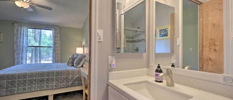 A bedroom with a bed, a ceiling fan, a window with curtains, and a bathroom with a sink and mirror.