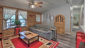 Ein Interieur einer Blockhütte mit Deckenventilator aus Holz, einem grauen Sofa, einem Holzschrank und einem roten Hocker.