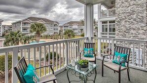 A balcony with two chairs and a table, overlooking a residential area with multiple houses and a stone wall. - Myrtlewood Golf Resort Condo With Pool Access (Myrtle Beach)