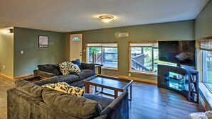 A spacious living room with a large sofa, a coffee table, and a flat-screen TV mounted on a wall. There is a sign indicating a guest area and a view of the outside through large windows. - Boat Dock, Game Room: Lakefront Sunrise Beach Home (Sunrise Beach)