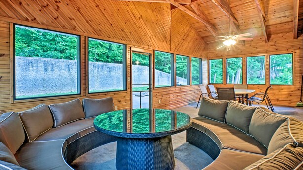 A wooden interior with a round glass table surrounded by cushioned seating, large windows with a view of greenery, and a ceiling fan. - Boat Dock, Game Room: Lakefront Sunrise Beach Home (Sunrise Beach)
