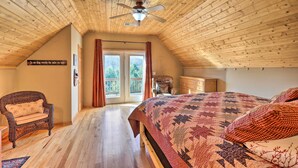 A cozy attic bedroom with a wooden ceiling, a large bed with a quilt, a wicker chair, and a view through a glass door to the outside.