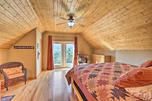 Un dormitorio acogedor en el ático, con techo de madera, una cama grande con colcha, una silla de mimbre y vistas al exterior a través de una puerta de cristal.