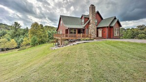 A large log cabin with a stone chimney, a wooden deck, and a grassy yard.