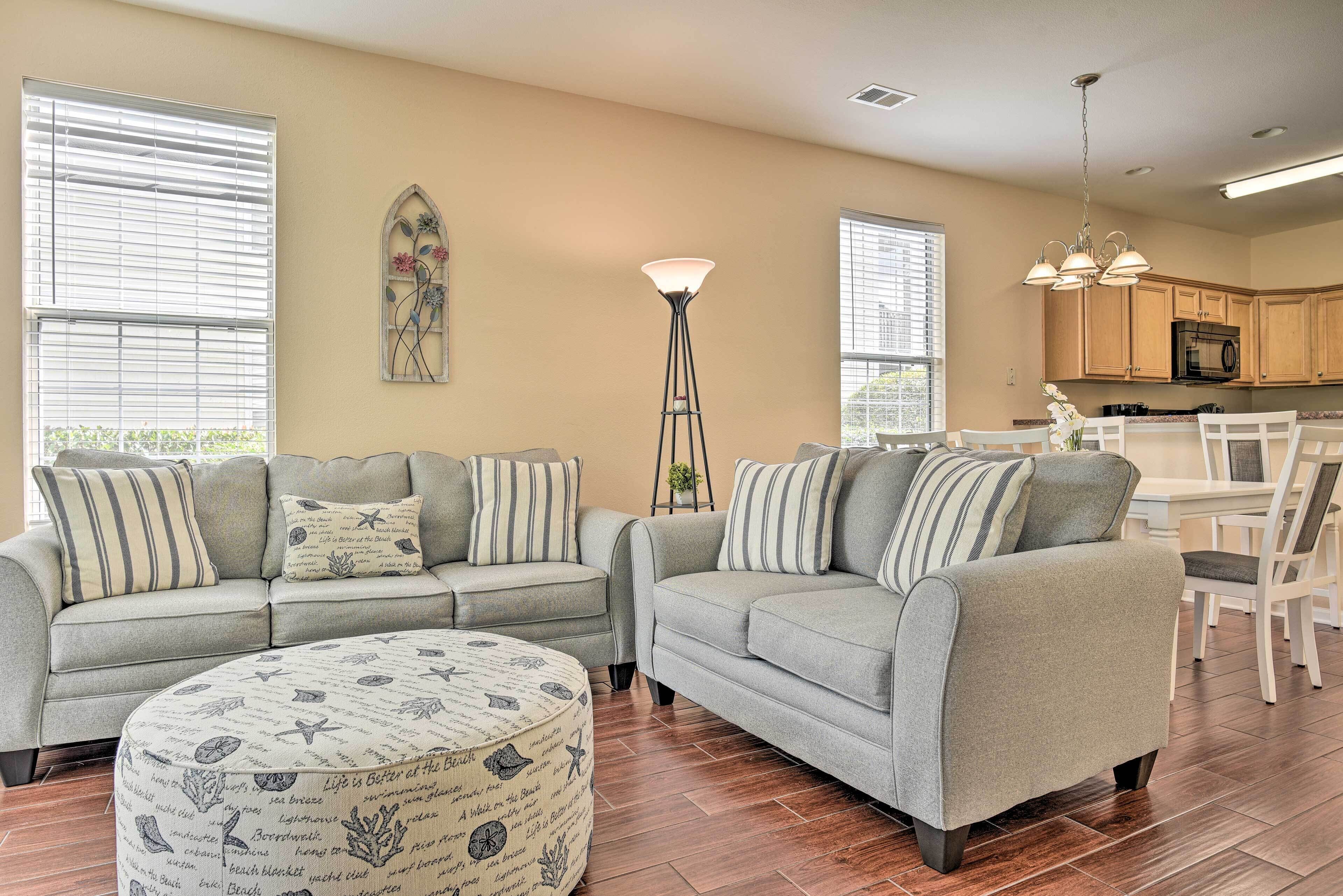 A living room with two grey sofas, a round ottoman, a tall floor lamp, and a kitchen in the background.