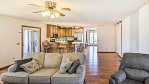 A modern living room with a sectional sofa, a gray armchair, and a coffee table. A kitchen with wooden cabinets and stainless steel appliances is visible in the background. - Country Home w/ Fire Pit: 40 Mi to Nashville! (Watertown)