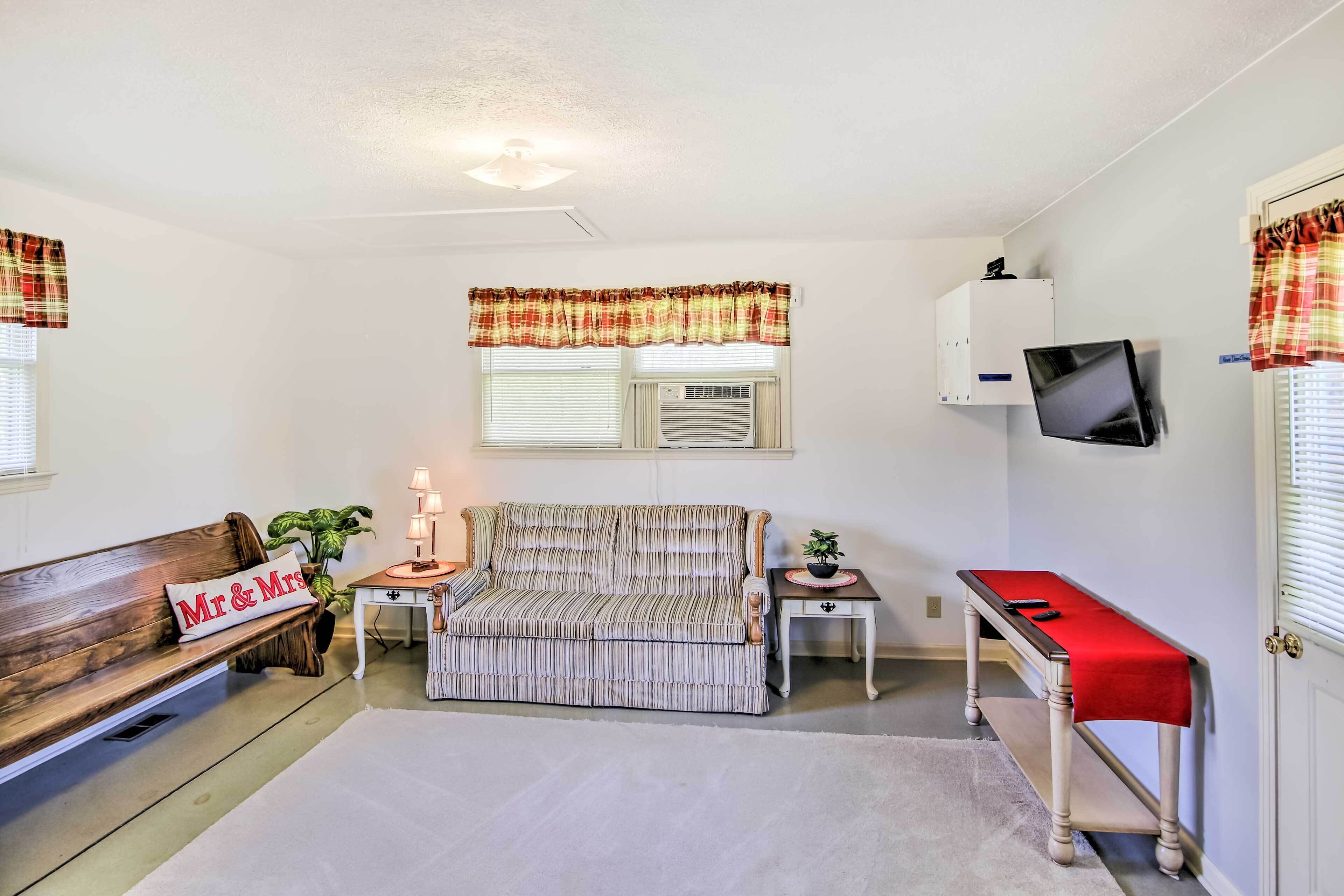 A cozy living room with a sofa, a bench, a small table with a red cloth, and a television mounted on the wall.