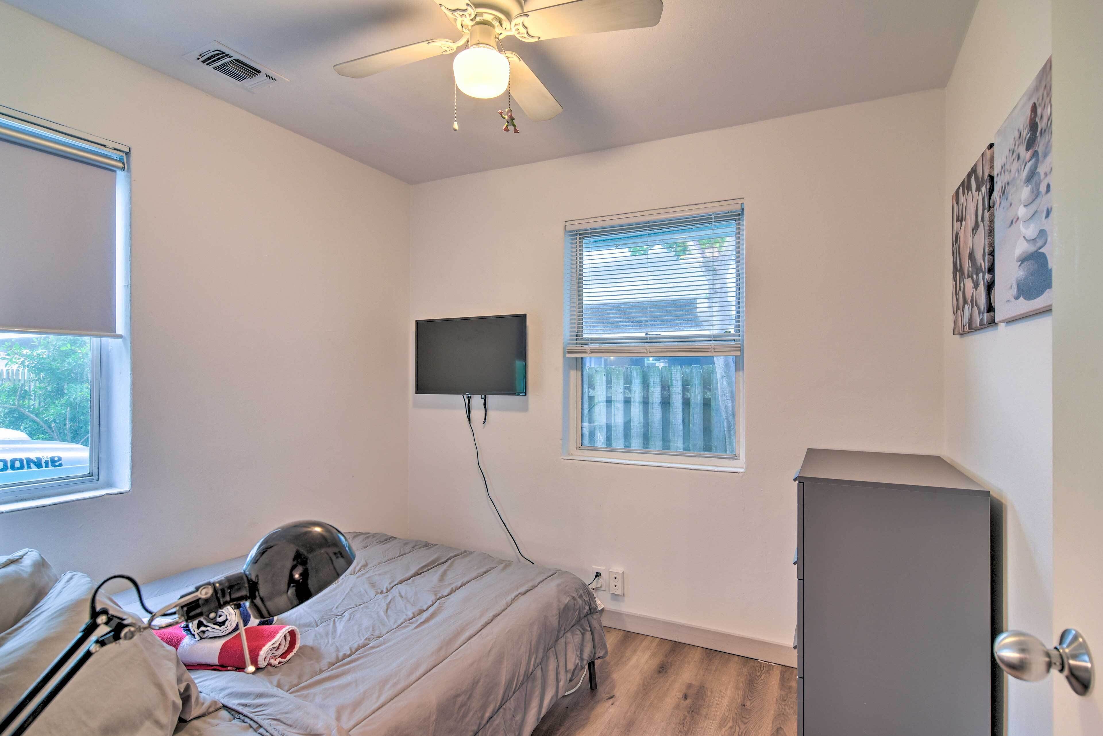 A bedroom with a bed, a ceiling fan, a TV mounted on the wall, a window with blinds, and a door.