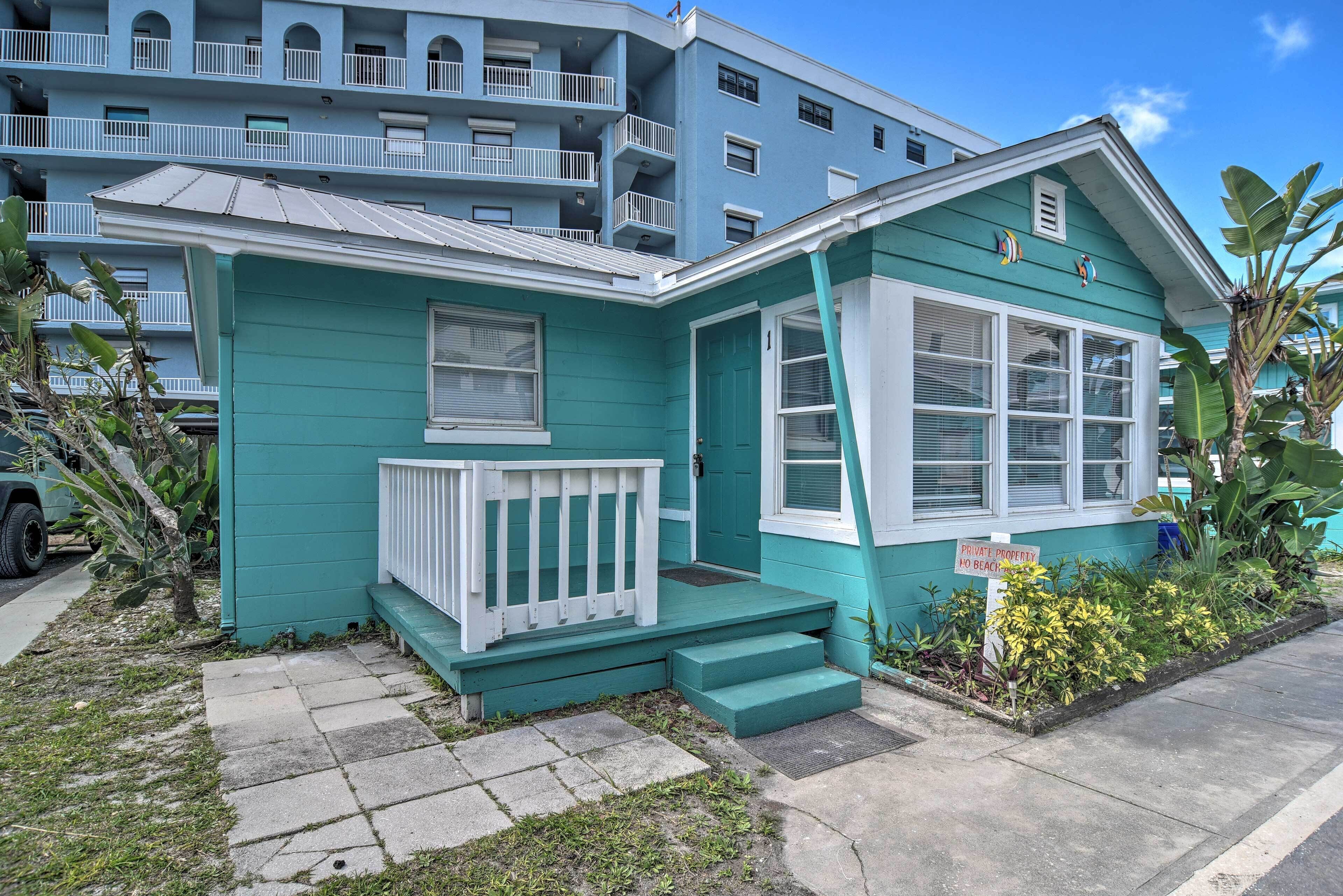 Une maison turquoise avec un porche blanc, un panneau indiquant « Propriété privée, accès à la plage interdit », et, au loin, une vue sur un immeuble à plusieurs étages.