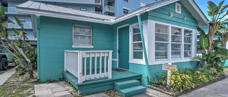 Une maison turquoise avec un porche blanc, un panneau indiquant « Propriété privée, accès à la plage interdit », et, au loin, une vue sur un immeuble à plusieurs étages.