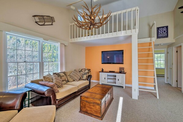 A spacious living room with a high ceiling, a large window, a cozy seating area, a wooden chest, and a white loft space. - Condo w/ Loft: 2 Mi to Windham Mountain Ski! (Windham)