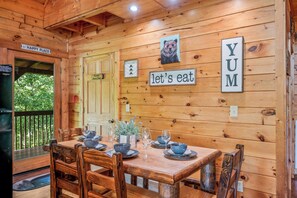 A wooden cabin interior with a dining table set for four, a bear picture, and signs saying "Happy Place" and "Let's eat YUM".