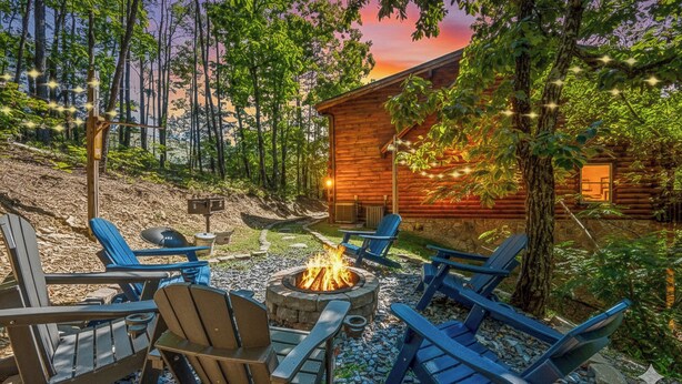 Una cabaña de troncos con un pozo de fuego rodeado de sillas Adirondack, ubicada en una zona boscosa.