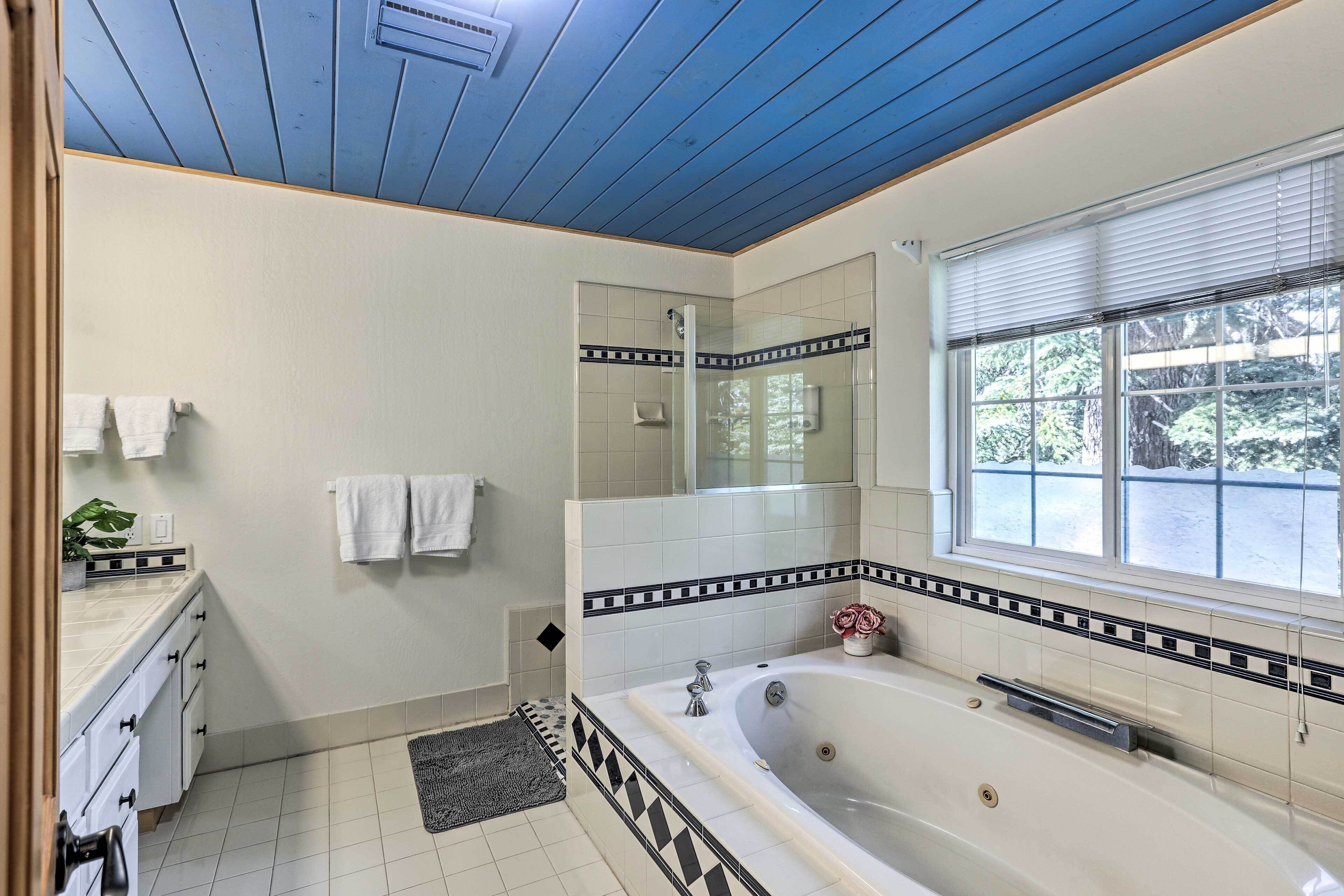A bathroom with a large bathtub, a window with blinds, a mirror, and a tiled floor.